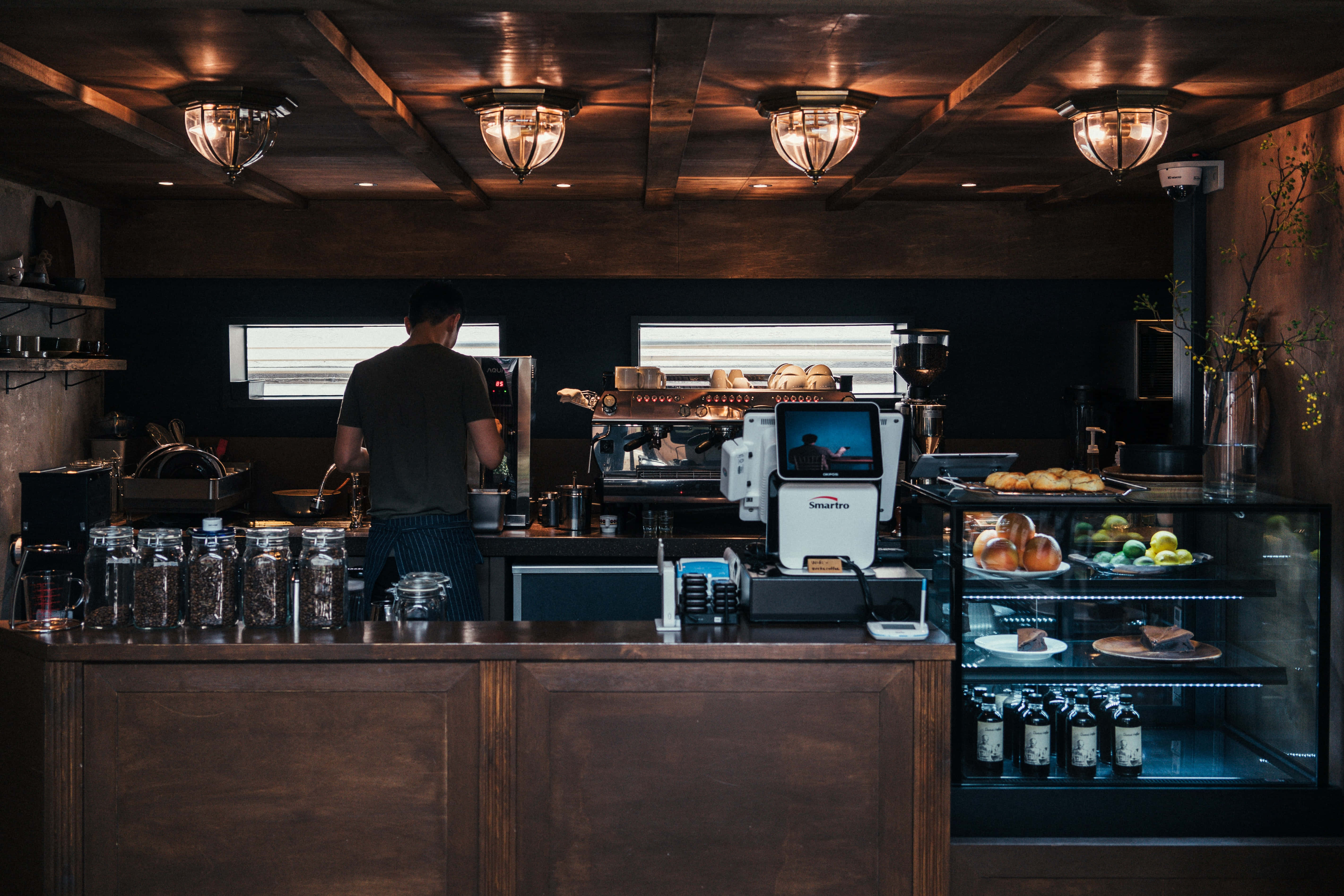 Coffee Aesthetic 桌布 壁紙 — 一位咖啡師在溫暖的吊燈下，在一個舒適昏暗的咖啡館裡，在深色木質咖啡櫃檯後工作。