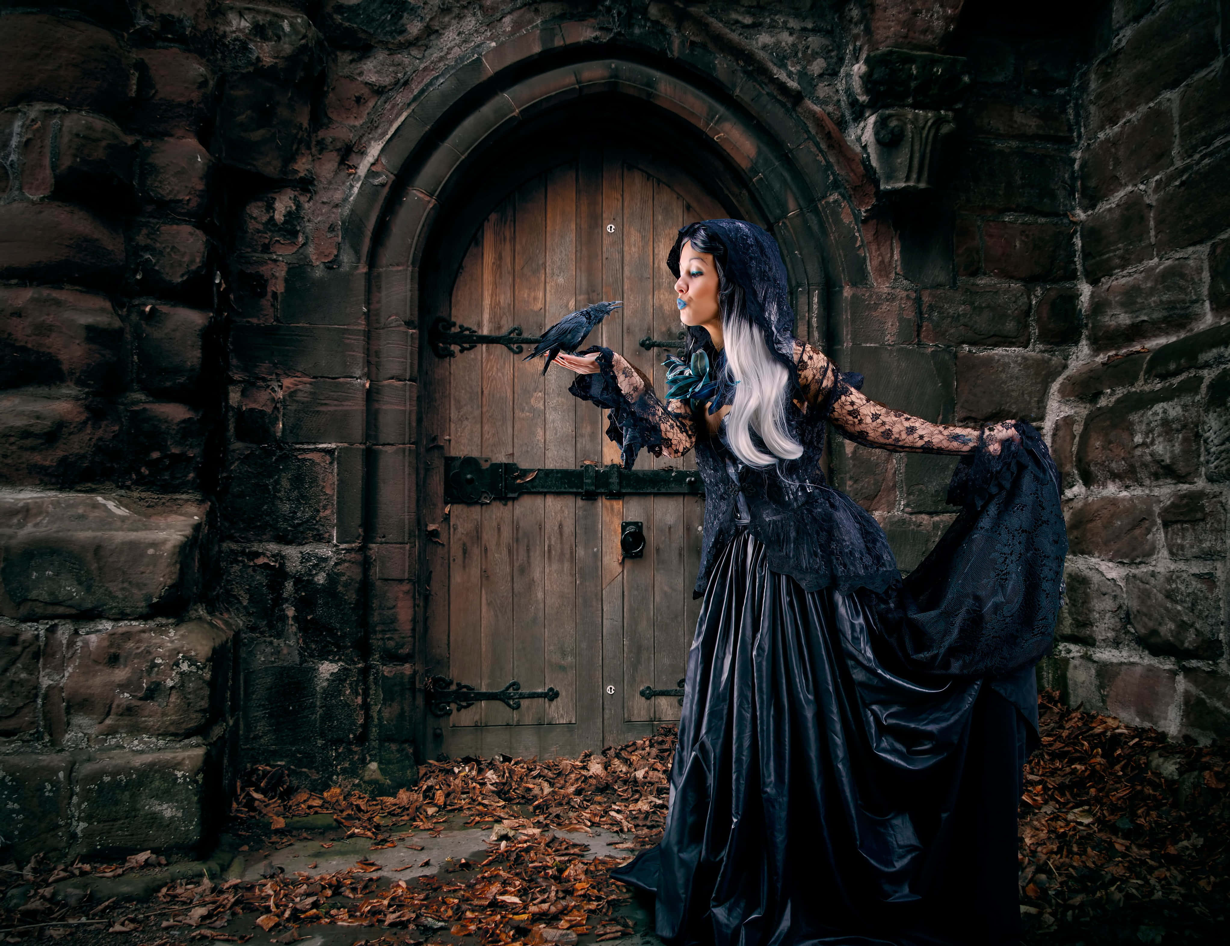 Gothic Costumes 桌布 壁紙 — 一位哥德式女子身穿黑色蕾絲長裙，站在風化的石拱門前，戴著手套的手上托著一隻烏鴉。