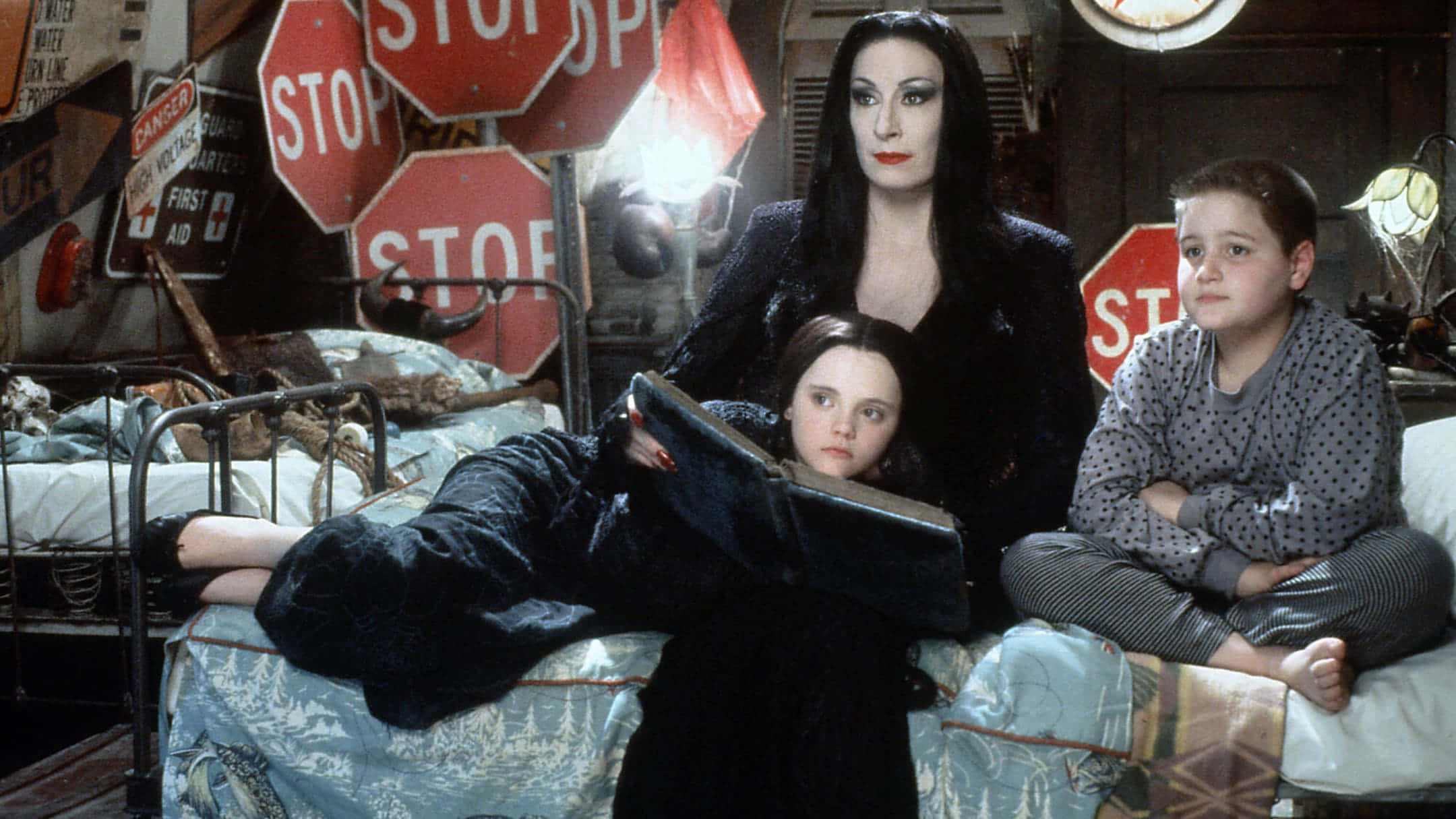 Addams Family 桌布 壁紙 — 一位哥德風格的母親和她的兩個孩子坐在床上，周圍環繞著停車標誌和陰森的裝飾，房間燈光昏暗。