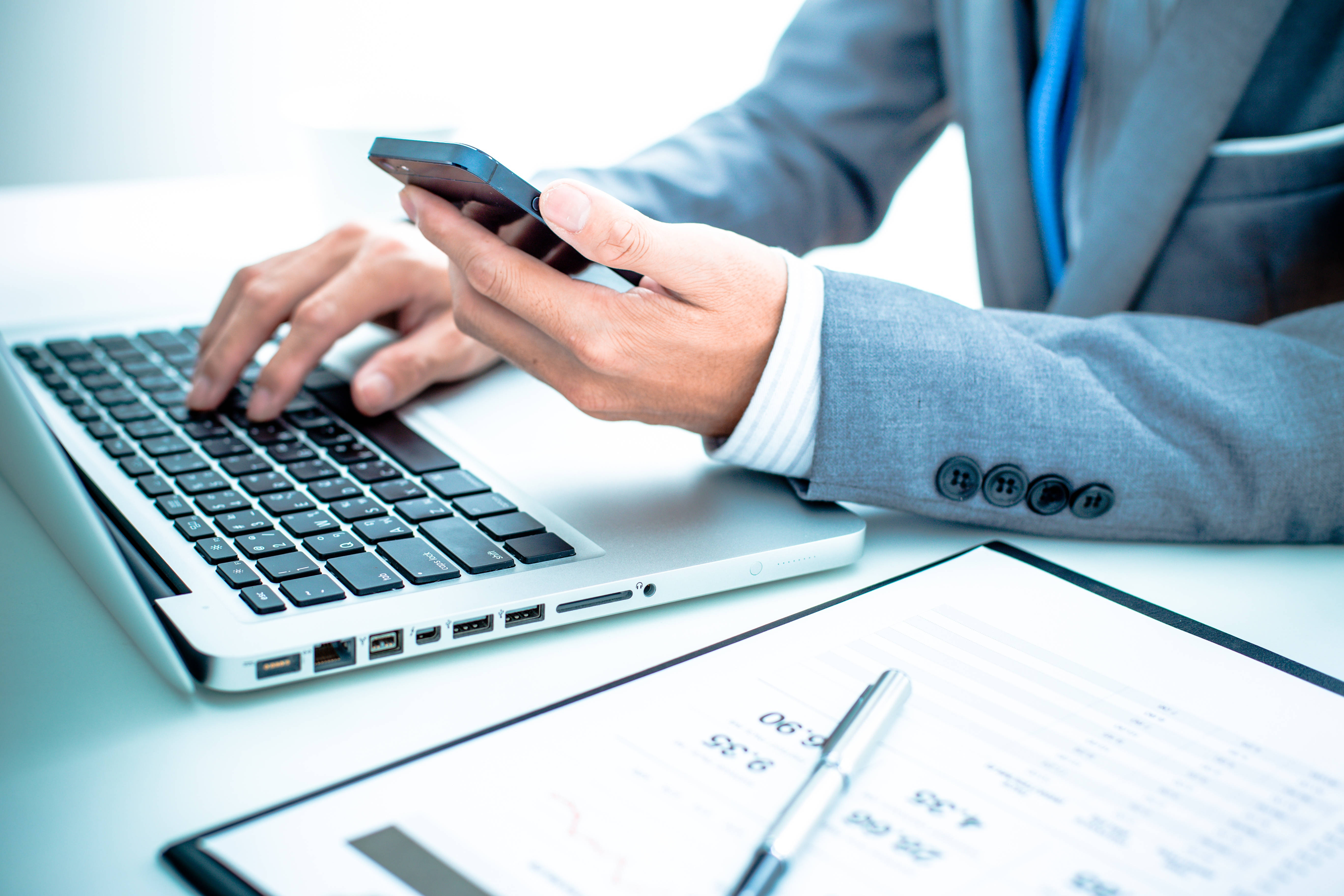 Chartered Accountant 桌布 壁紙 — 一位商人在辦公桌上使用筆記型電腦和智慧型手機同時處理多項任務，並審閱文件。