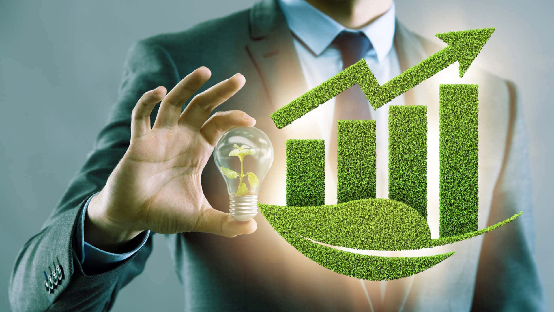 Green Economy 桌布 壁紙 — 一位商人手持一個發光的燈泡，裡面有嫩芽，旁邊漂浮著一個草質的上升條形圖和一片葉子。
