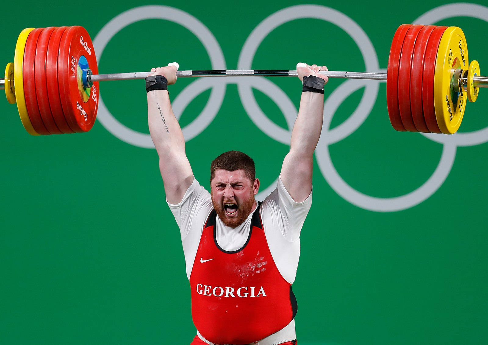 Olympics 桌布 壁紙 — 一位喬治亞舉重運動員在綠色奧運背景下，以堅毅和力量將沉重的槓鈴舉過頭頂。