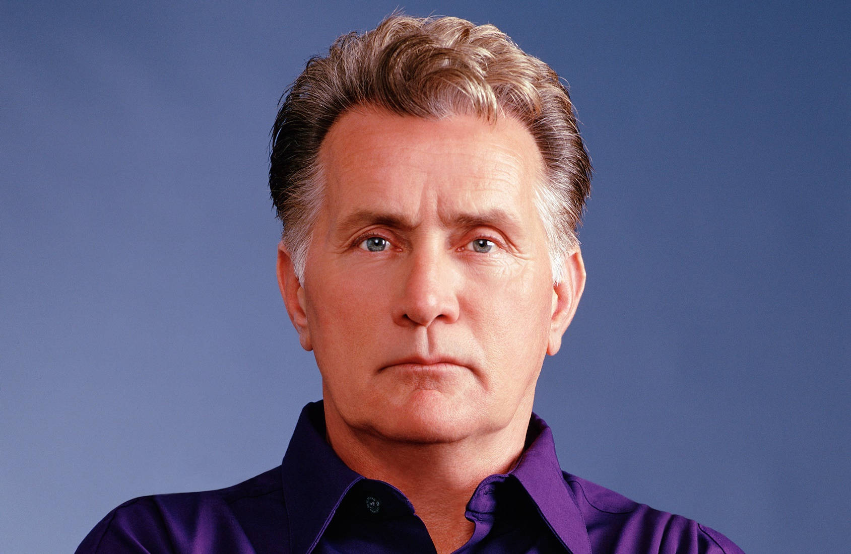actor 桌布 壁紙 — 一位嚴肅的中年男子特寫肖像，灰白頭髮，穿著紫色襯衫，背景為純藍色。