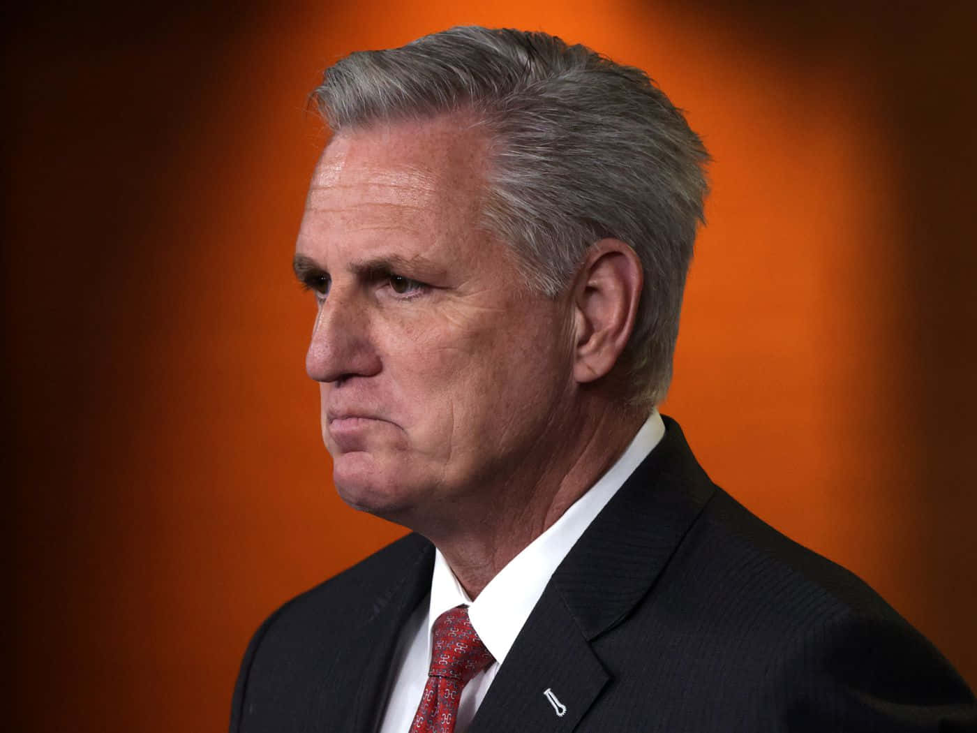 Politics 桌布 壁紙 — 一位嚴肅的中年男子身穿深色西裝和紅色領帶，在溫暖的橙色漸變背景前凝視著。