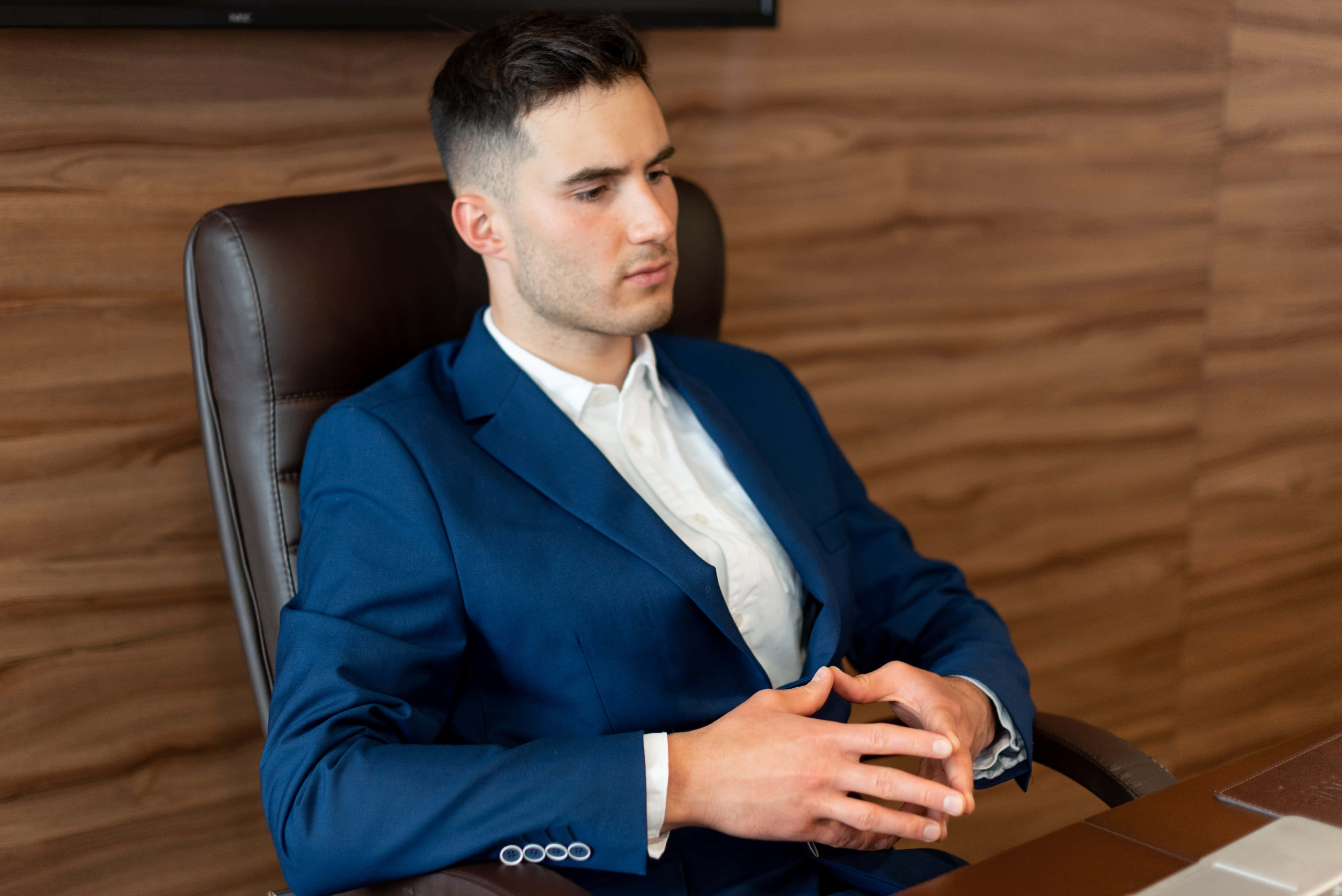 Navy Blue Suit 桌布 壁紙 — 一位嚴肅的商人穿著藍色西裝，坐在皮椅上，靠著木質鑲板的辦公室牆壁沉思。