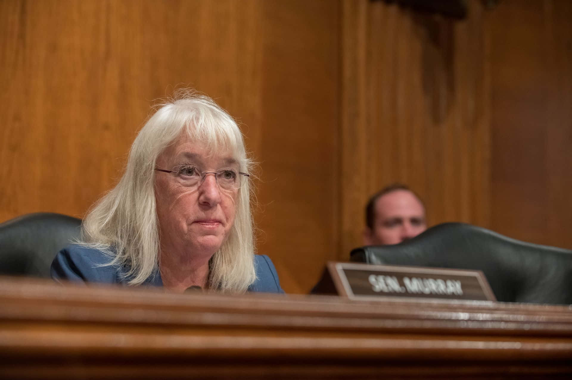 Senators 桌布 壁紙 — 一位嚴肅的白髮戴眼鏡女性坐在正式聽證室的木桌旁，桌上名牌寫著SEN MURRAY。