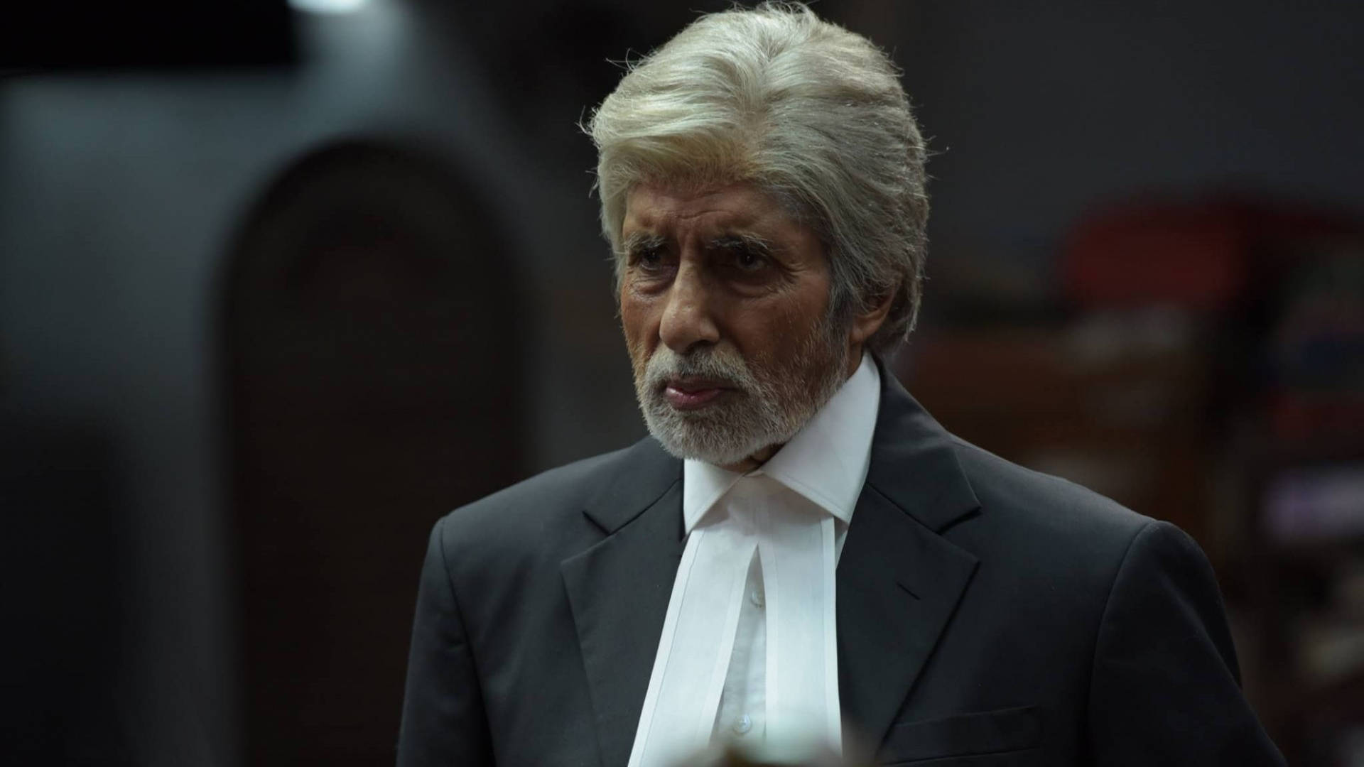 Amitabh Bachchan 桌布 壁紙 — 一位嚴肅的白髮白鬍鬚老年男子，穿著黑色西裝和白色法官袍，背景是燈光昏暗的法庭。