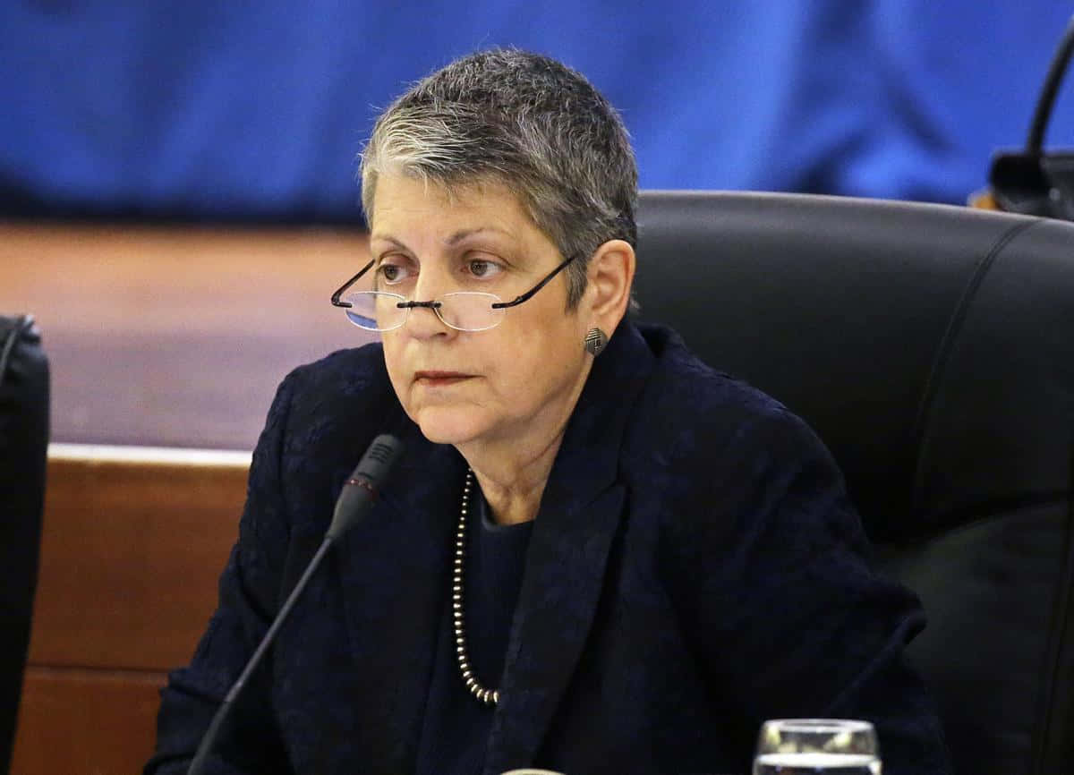 政治 桌布 壁紙 — 一位嚴肅的短灰髮戴眼鏡女士坐在藍色背景的正式會議室的木桌旁。