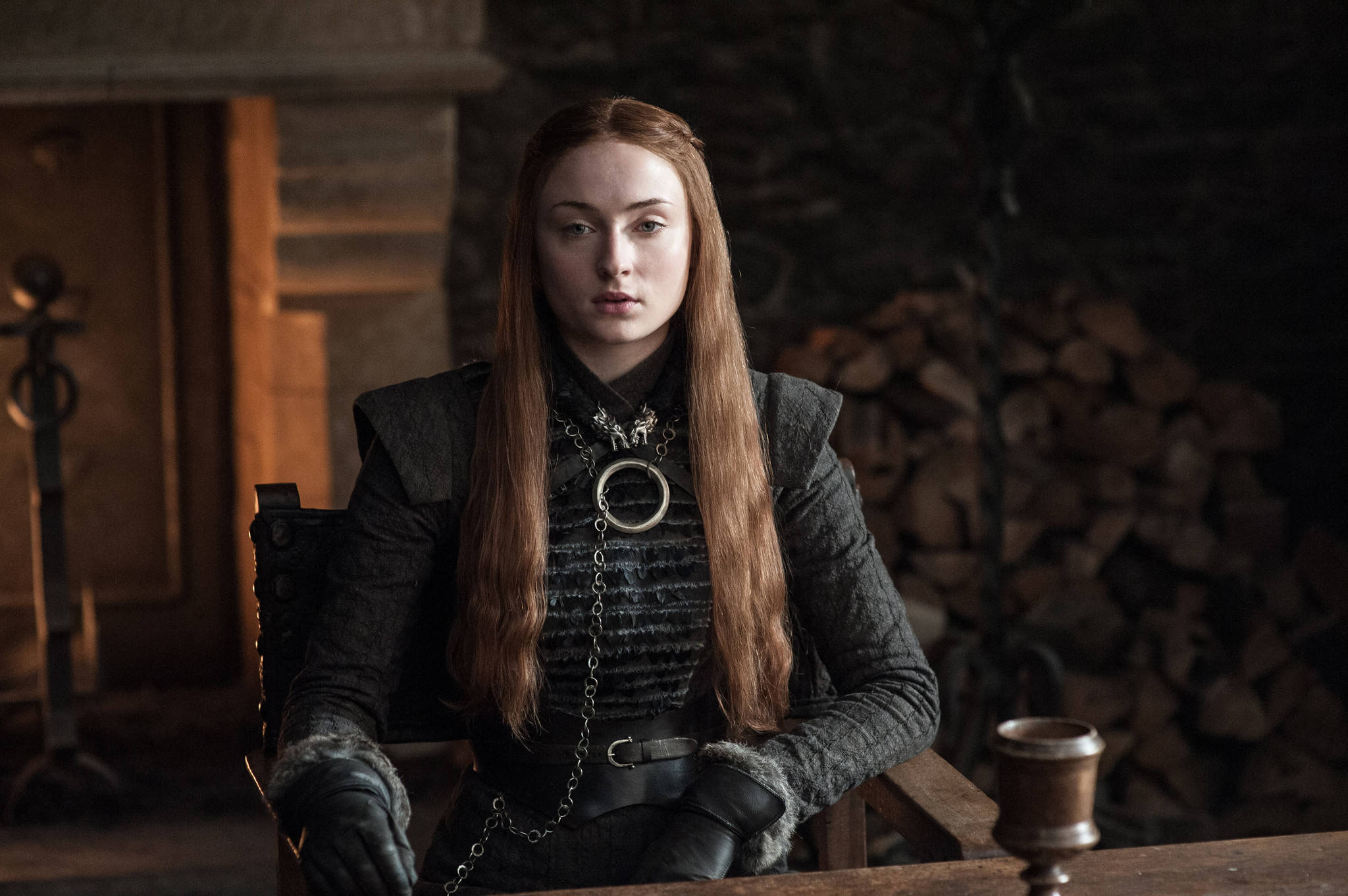 Sansa Stark 桌布 壁紙 — 一位嚴肅的長紅髮女性坐在光線昏暗的中世紀房間裡，身後有石壁爐和堆疊的木柴。