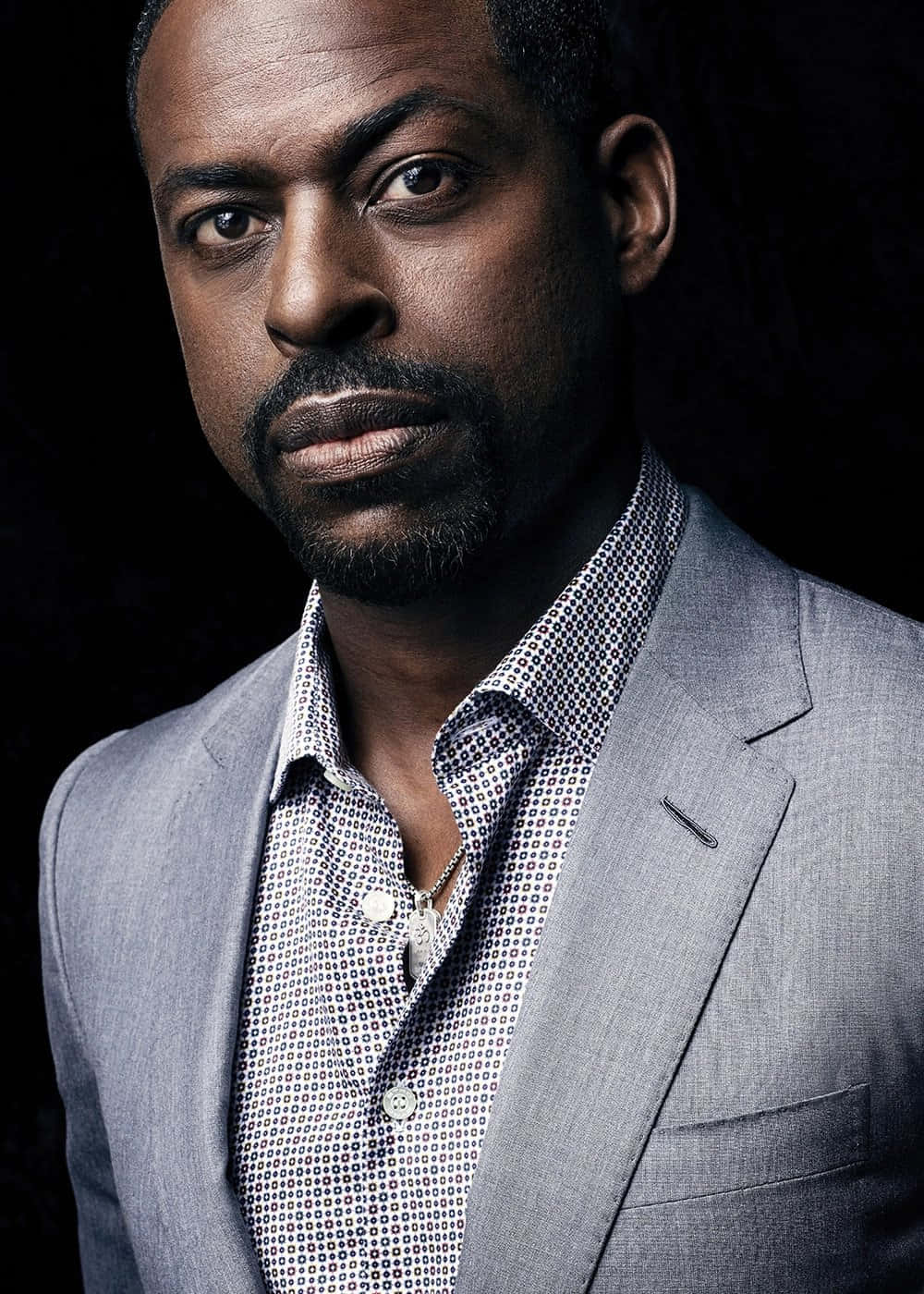 Sterling K Brown 桌布 壁紙 — 一位嚴肅的黑人男子身穿灰色西裝和圖案襯衫，在深色背景下強烈凝視。