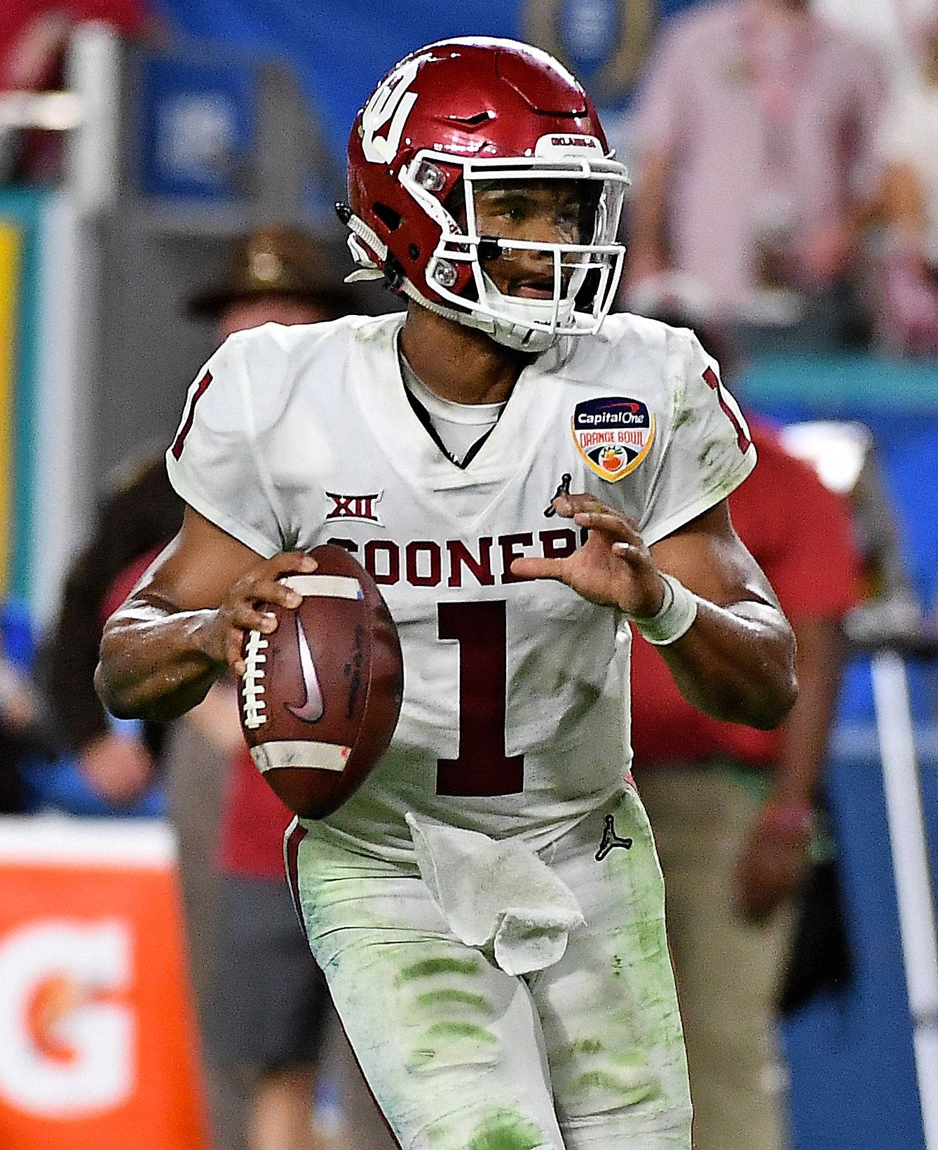 運動 桌布 壁紙 — 一位堅定的奧克拉荷馬大學 Sooners 美式足球四分衛，身穿白色和深紅色裝備，在體育館燈光下的比賽中帶球奔跑。