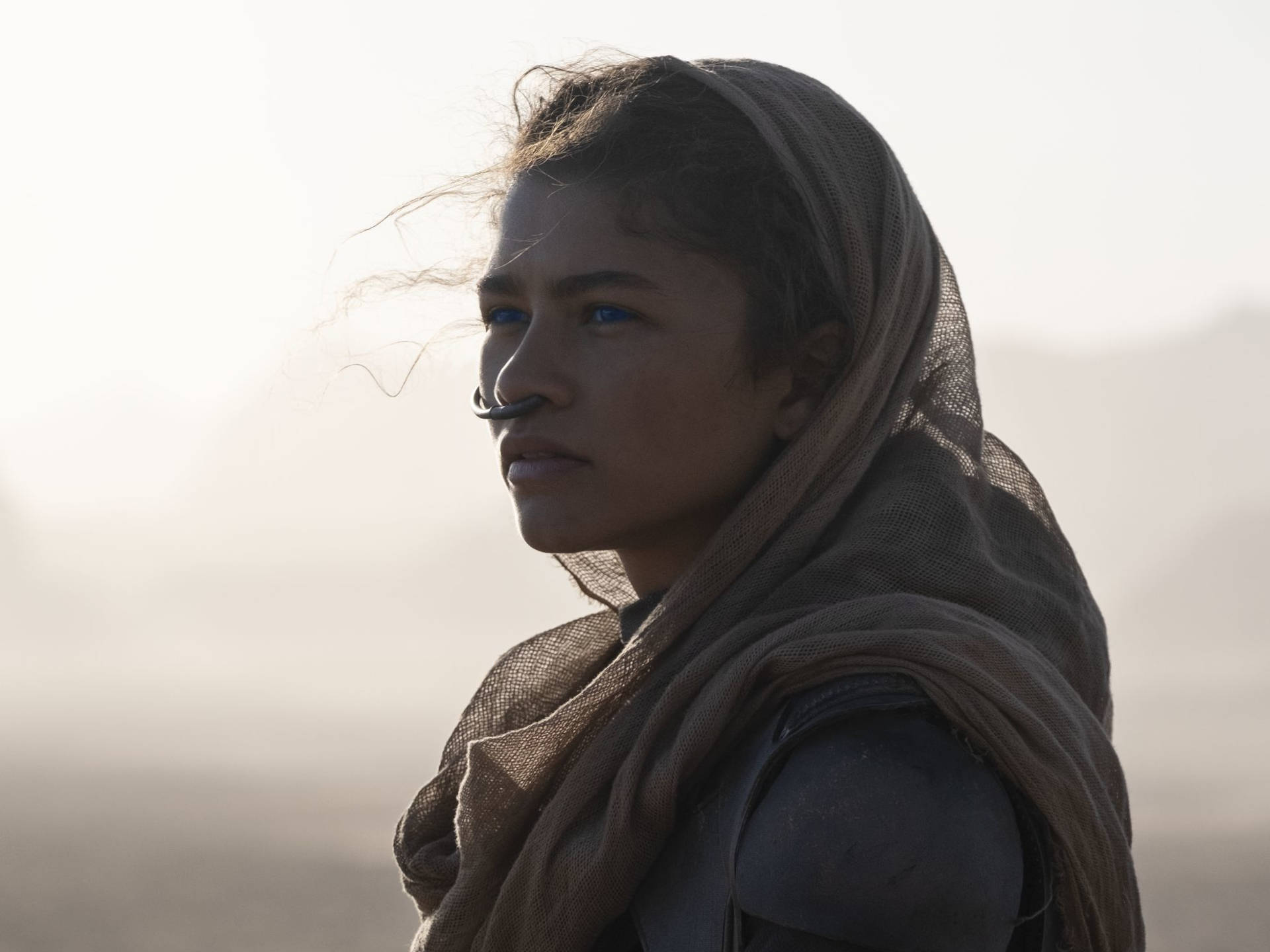 Dune 2021 桌布 壁紙 — 一位堅定的女性，藍眼睛，戴著頭巾，站在日出時朦朧的沙漠景觀中。