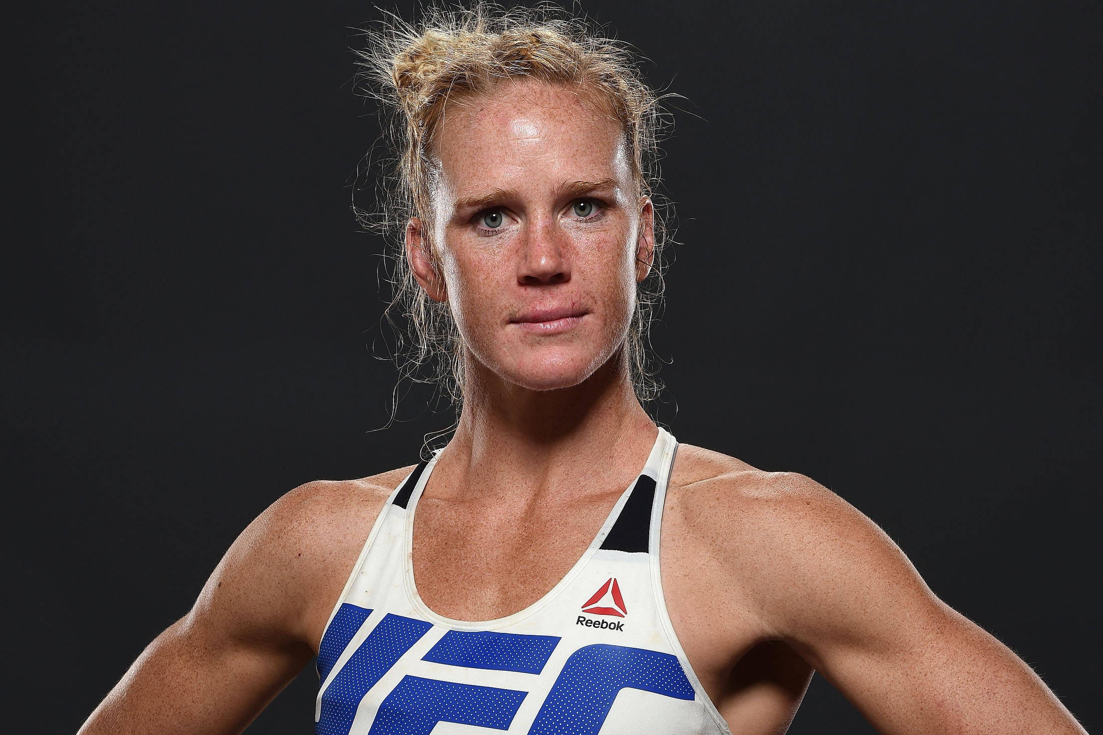 MMA 桌布 壁紙 — 一位堅定的女性MMA拳手，身穿白色和藍色的Reebok UFC上衣，在深色背景下凝視著。