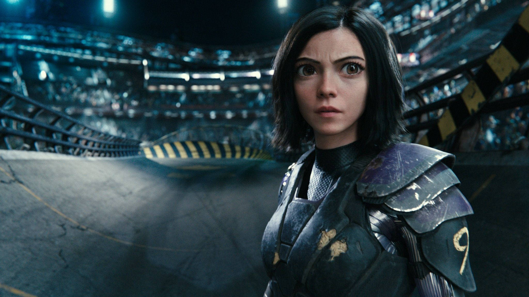 Alita Battle Angel 桌布 壁紙 — 一位堅定的女戰士穿著未來風格的盔甲，站在燈光昏暗的競技場中，周圍有發光的燈光和模糊的觀眾。