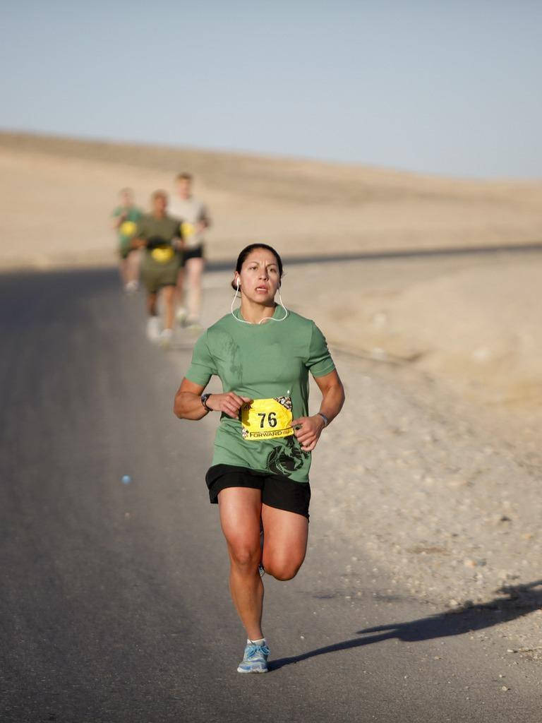 Marathon 桌布 壁紙 — 一位堅定的女跑者，身穿綠色襯衫和黑色短褲，在陽光明媚的沙漠道路上衝刺，其他跑者緊隨其後。