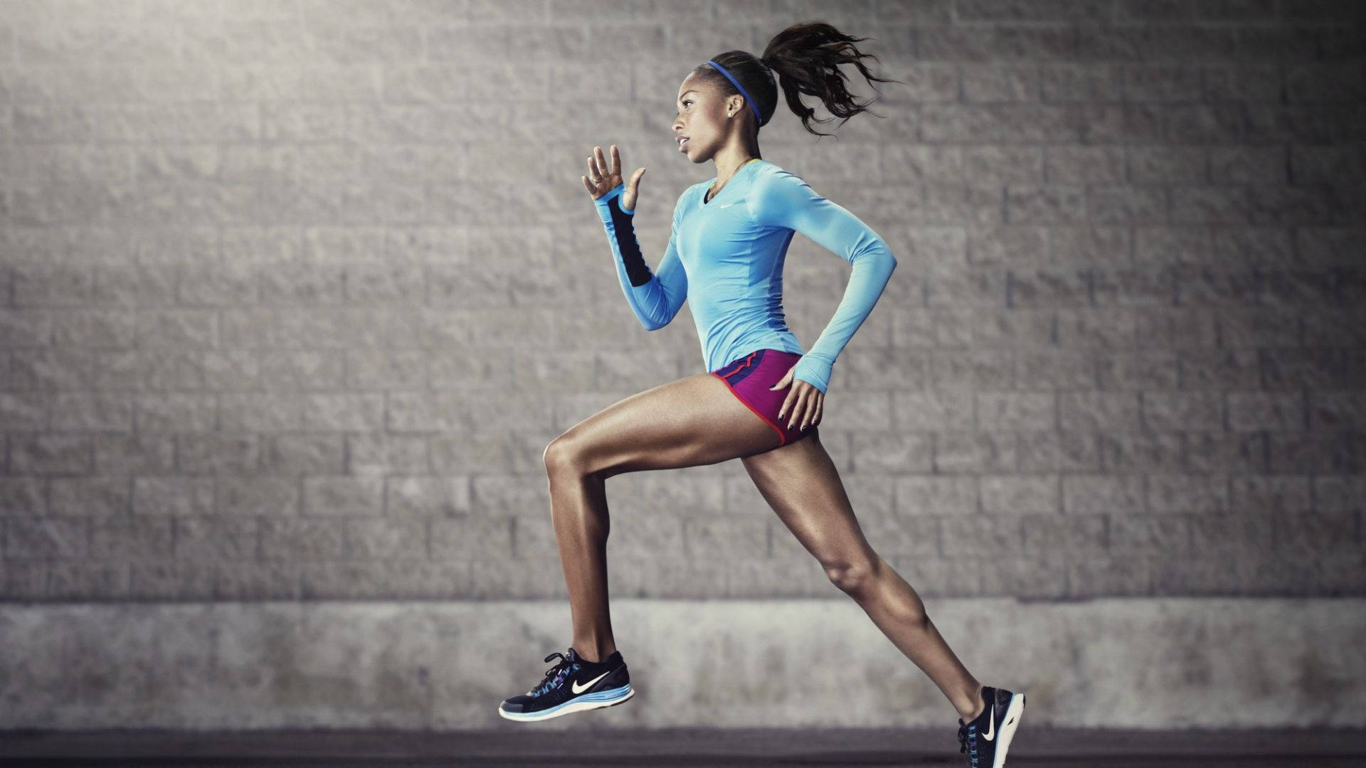 nike girl 桌布 壁紙 — 一位堅定的女跑者，身穿藍色上衣和洋紅色短褲，以動態動作衝過灰色磚牆。
