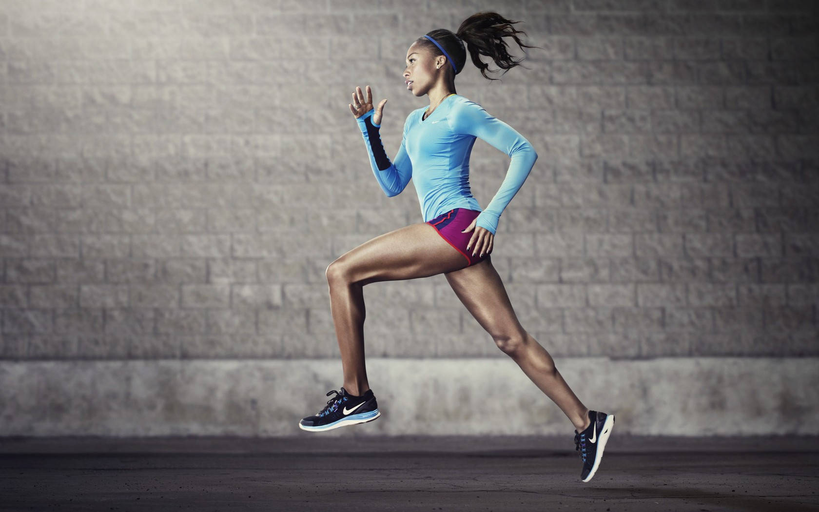 Exercise 桌布 壁紙 — 一位堅定的女跑者身穿藍色上衣和紫色短褲，在帶有紋理的灰色磚牆前以中步幅衝刺。