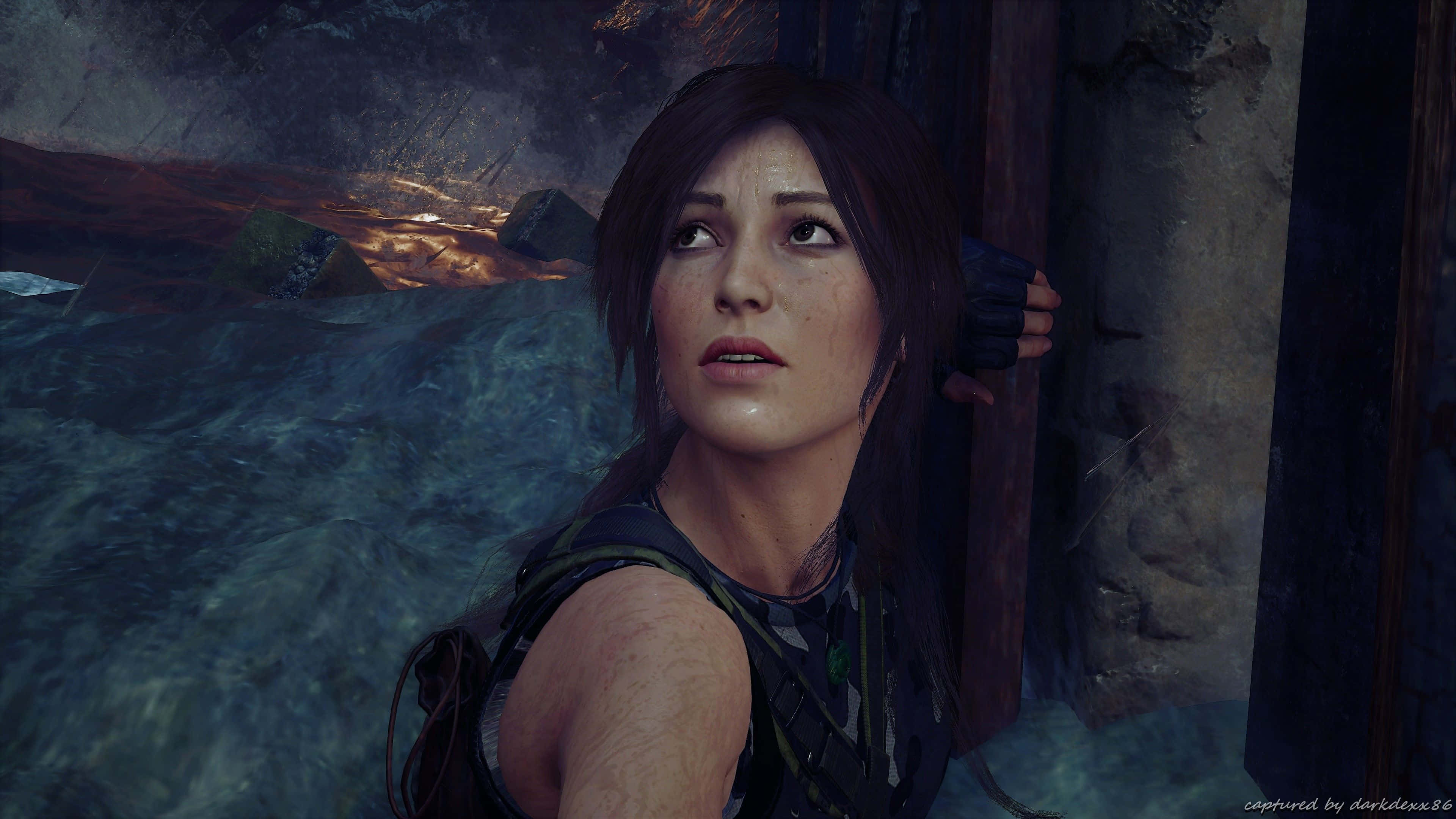 Lara Croft 桌布 壁紙 — 一位堅定的探險家，深色頭髮，穿著堅固裝備，站在石柱旁，在昏暗的廢墟中仰望，背景有發光的橙色餘燼。