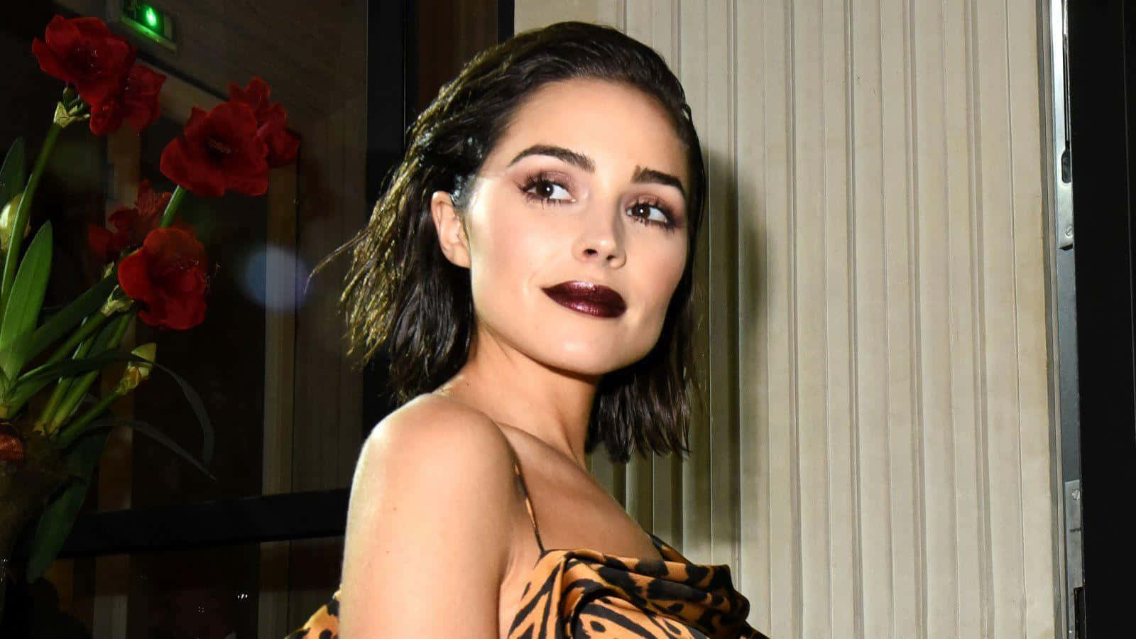 olivia culpo 桌布 壁紙 — 一位塗著深色唇膏、短髮的女性，身穿豹紋上衣，在紅花和垂直百葉窗旁擺姿勢。