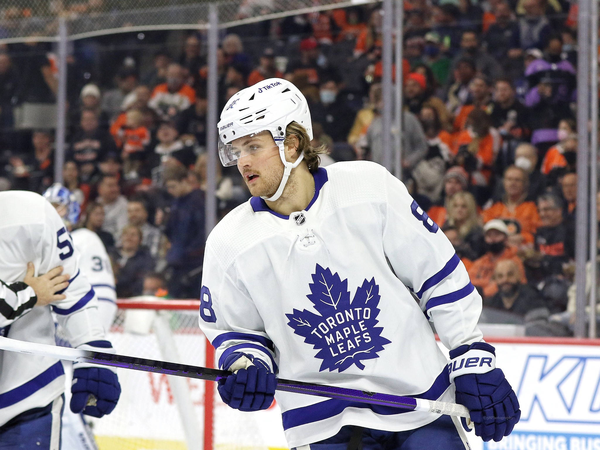 hockey 桌布 壁紙 — 一位多倫多楓葉隊的冰球運動員身穿白色和藍色裝備，站在冰上手持球桿，背景中穿著橙色球衣的球迷模糊不清。