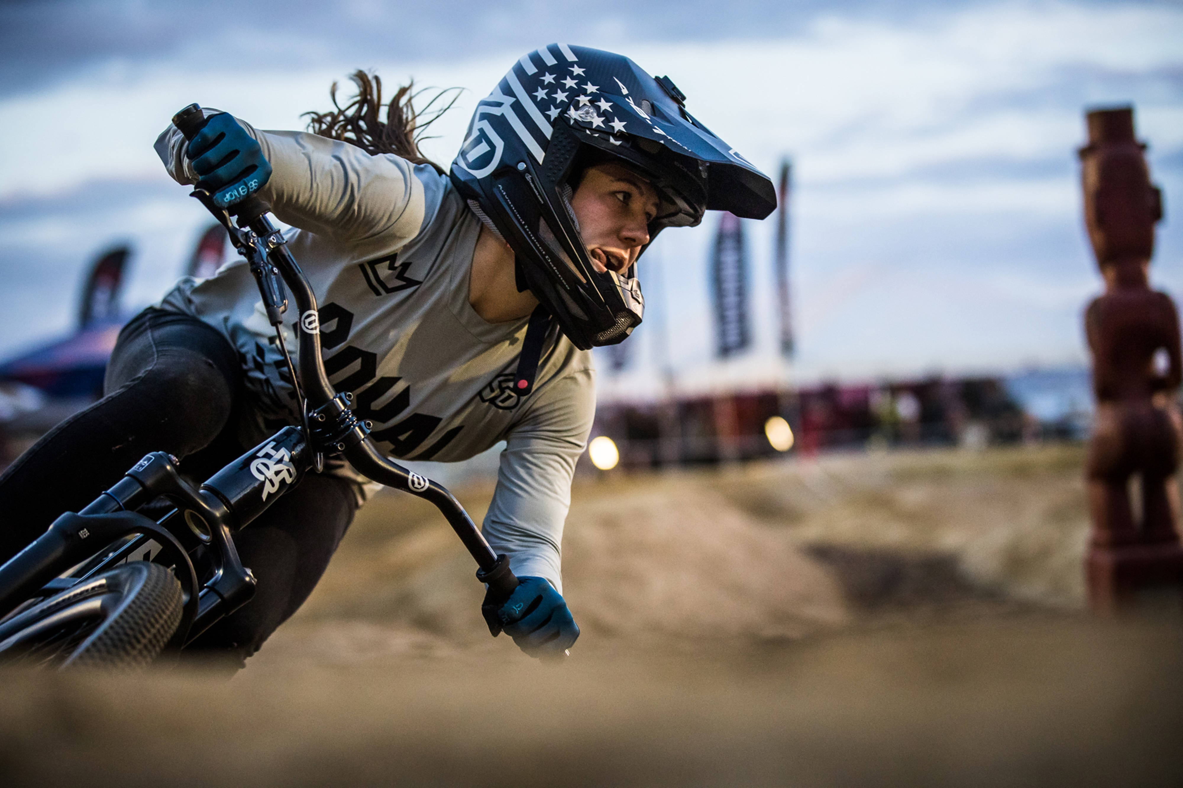 Woman 桌布 壁紙 — 一位女BMX車手在泥地賽道上傾身過急彎，戴著頭盔，神情專注。