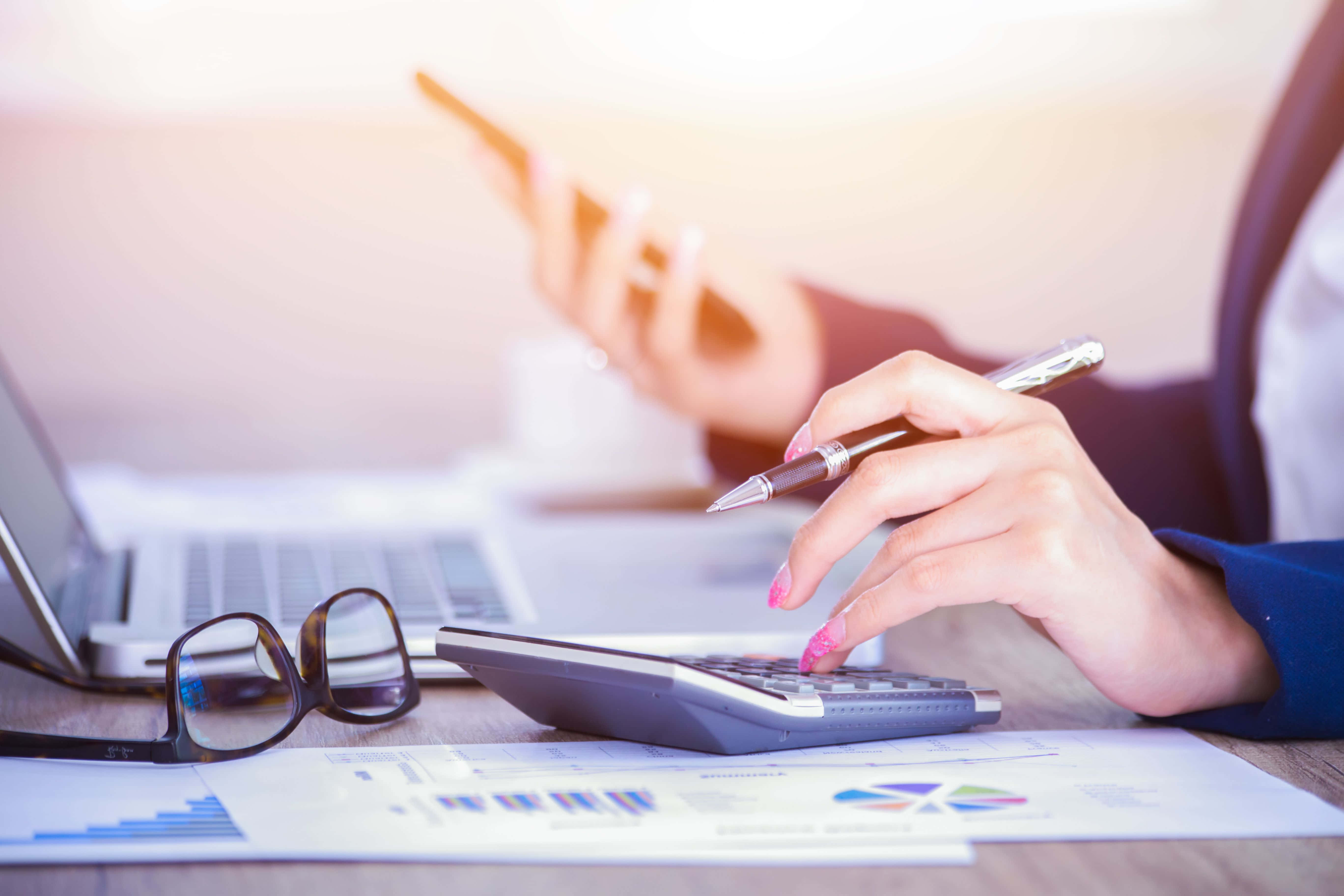 Accounting 桌布 壁紙 — 一位女商人的手在使用計算機和智慧型手機，同時在木桌上審閱財務圖表。