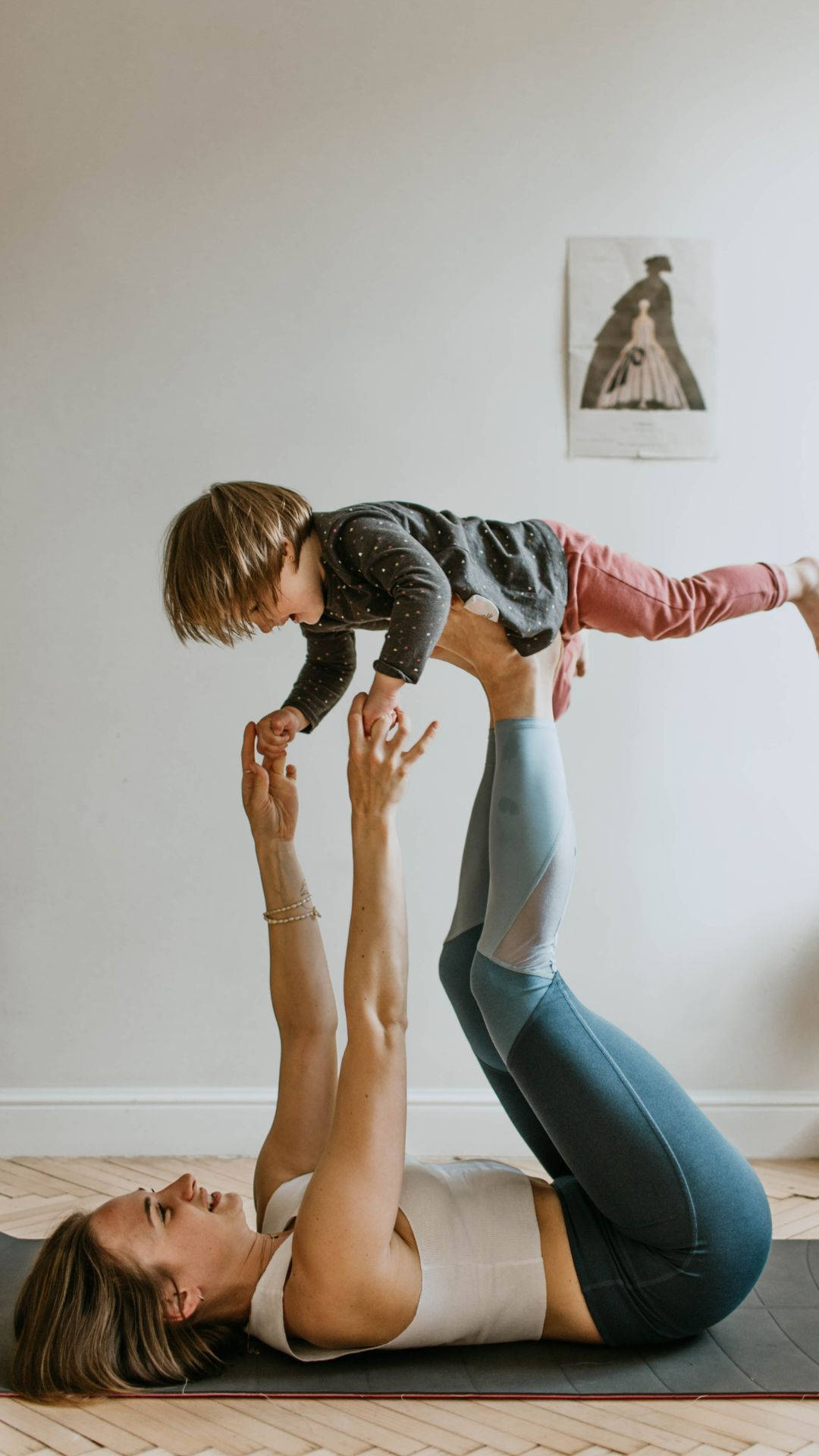 Mom 桌布 壁紙 — 一位女士以瑜伽姿勢支撐著一個在她腳上保持平衡的小孩，身處明亮簡約的房間。