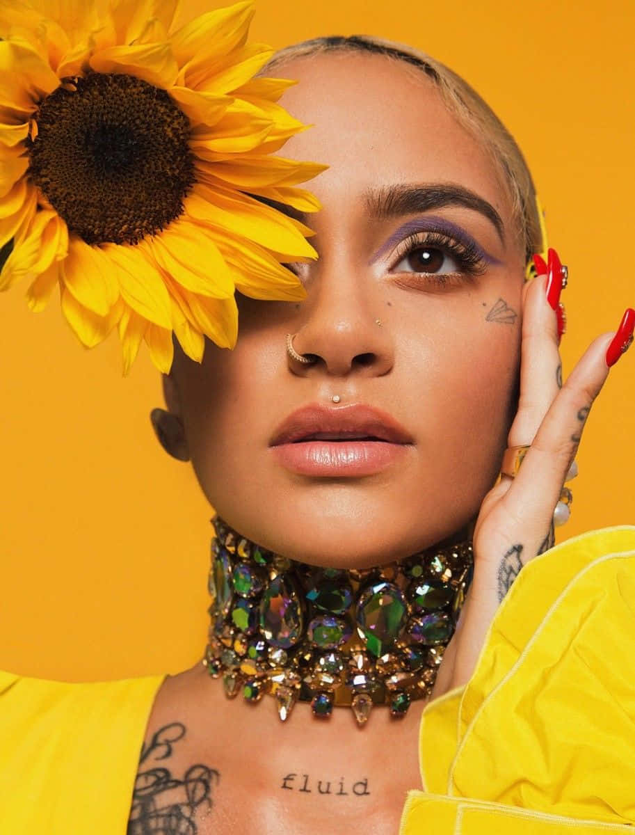 kehlani 桌布 壁紙 — 一位女士化著大膽的紫色眼妝，眼睛上放著一朵大向日葵，在鮮黃色背景前擺姿勢。