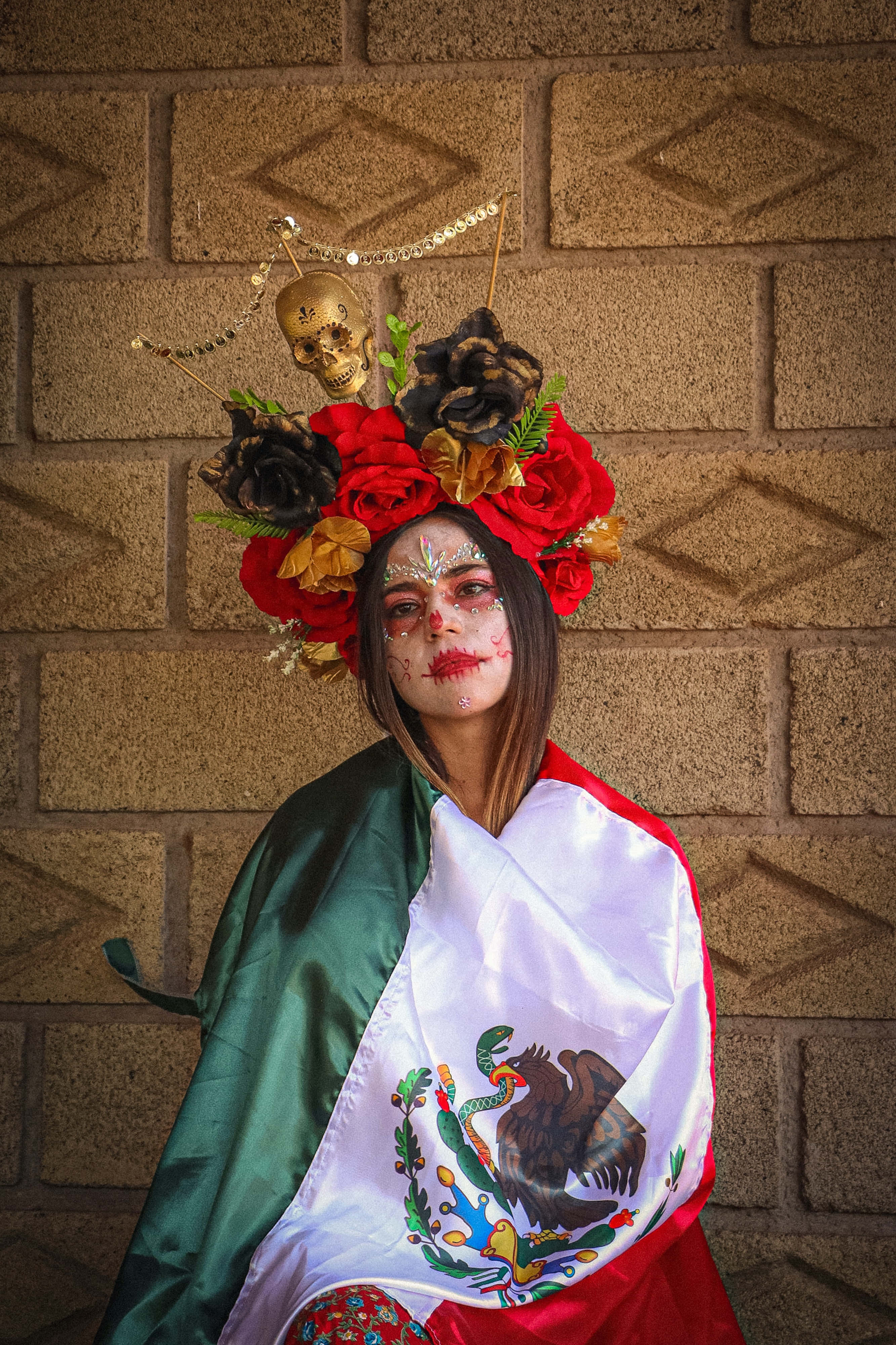 Day Of The Dead 桌布 壁紙 — 一位女士化著鮮豔的糖骷髏妝容，戴著花卉頭飾，披著墨西哥國旗，站在磚牆前。
