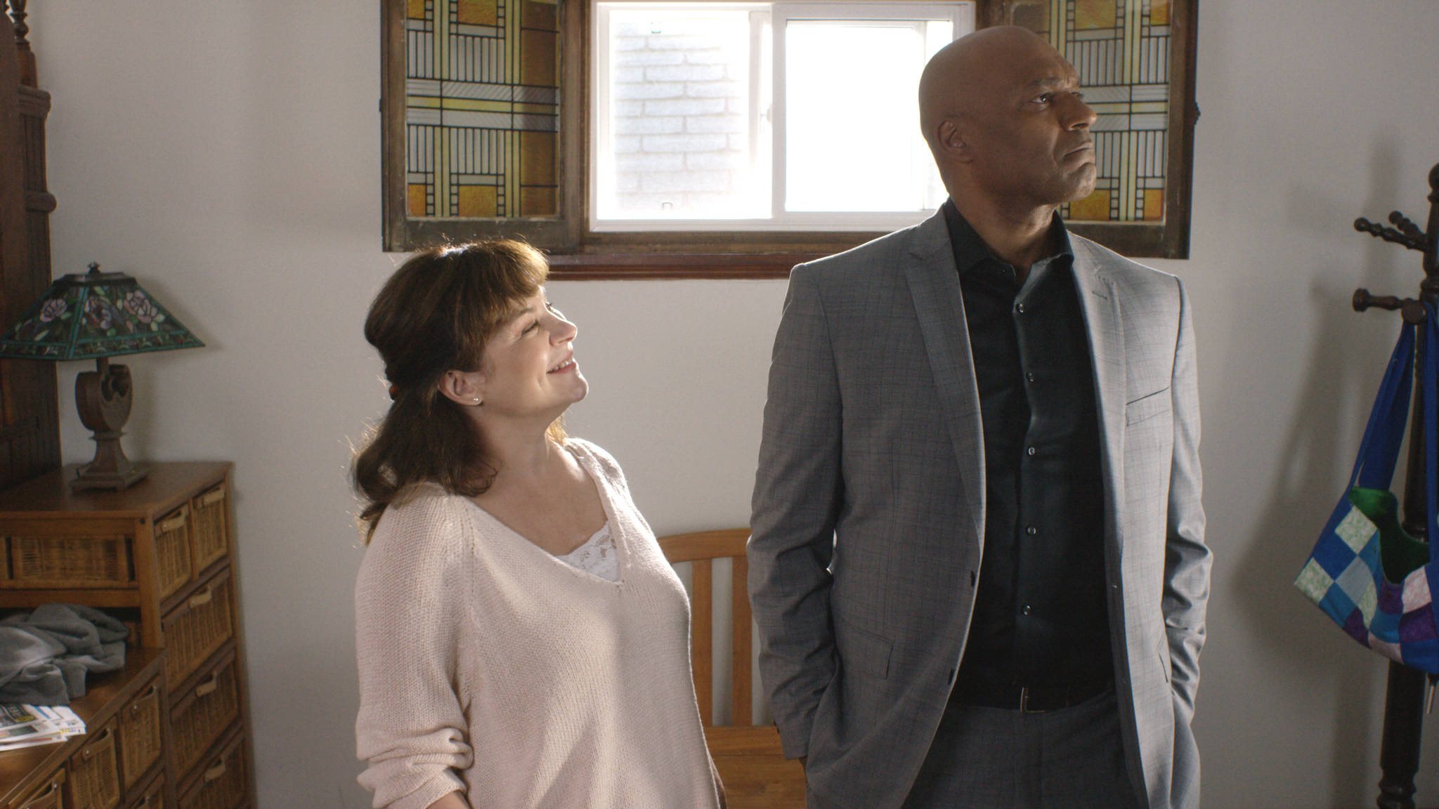 Colin Salmon 桌布 壁紙 — 一位女士向一位身穿灰色西裝、站在有彩繪玻璃窗的溫暖燈光房間裡的男士微笑。