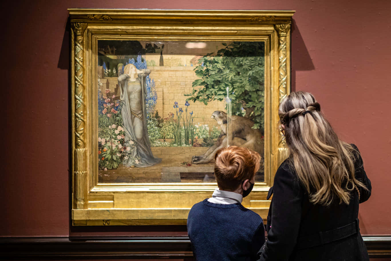 Museum 桌布 壁紙 — 一位女士和一個孩子在博物館透過金色畫框觀看一幅古典繪畫，畫中描繪了一個披著布的人物和一隻猴子在花園裡。