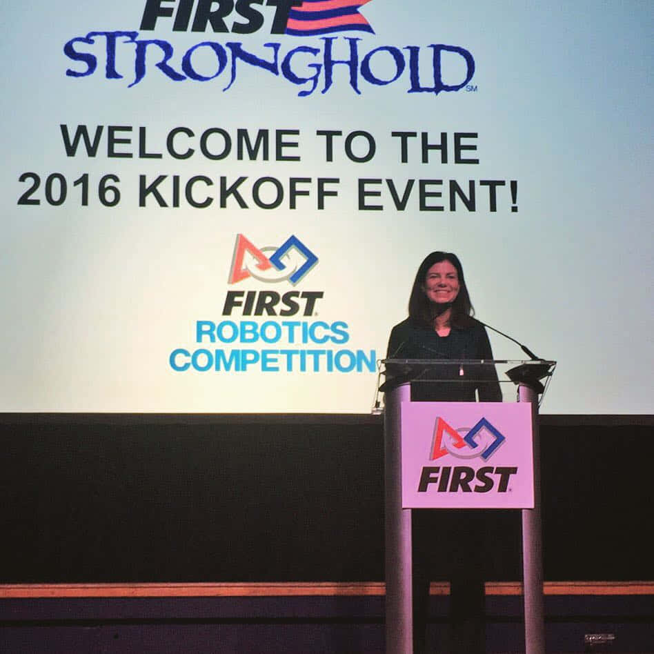 政治 桌布 壁紙 — 一位女士在2016年FIRST Robotics Competition啟動活動中在講台上微笑，身後有品牌標誌。