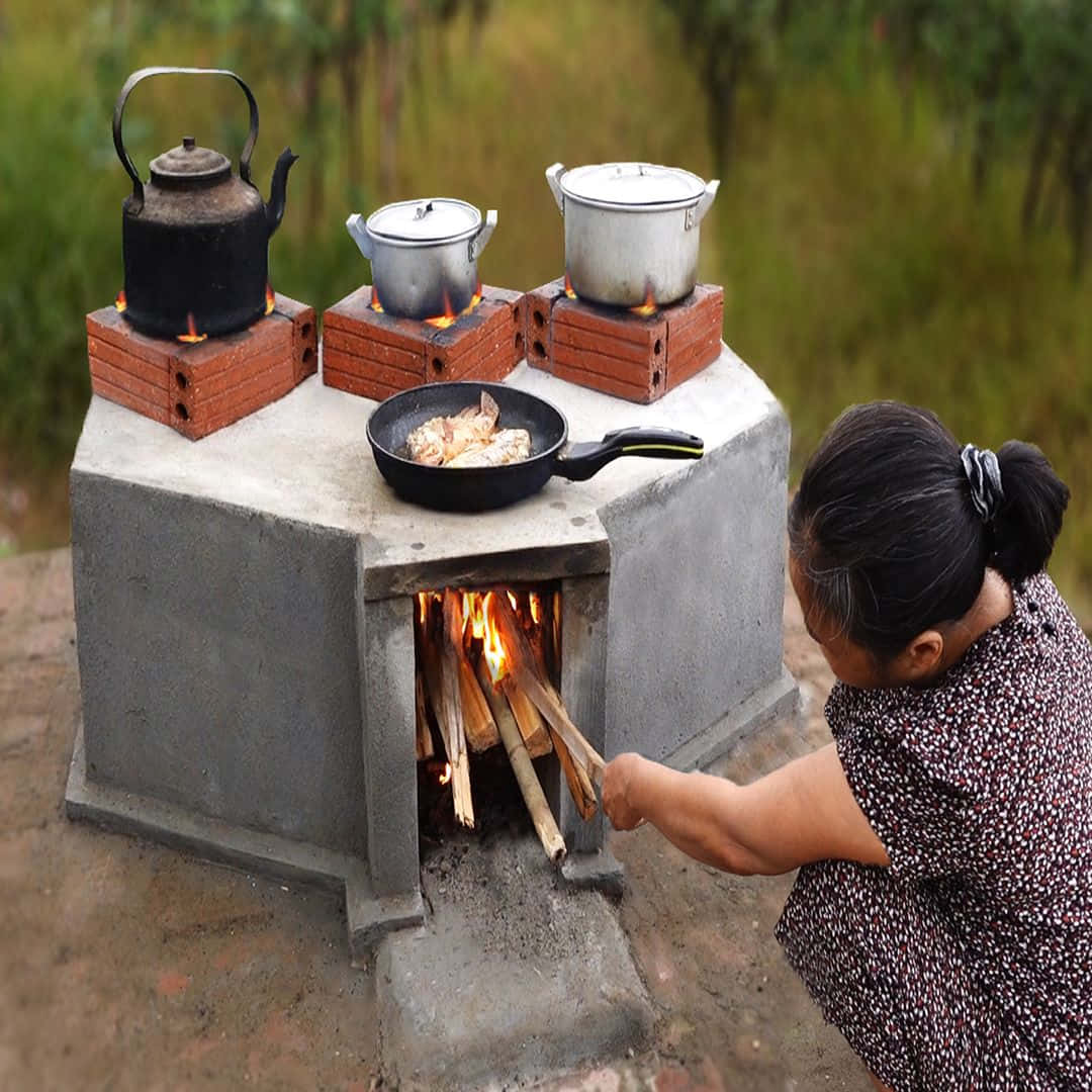 Stove 桌布 壁紙 — 一位女士在戶外照料一個燒柴的爐灶，鍋子和平底鍋在明火上慢煮。