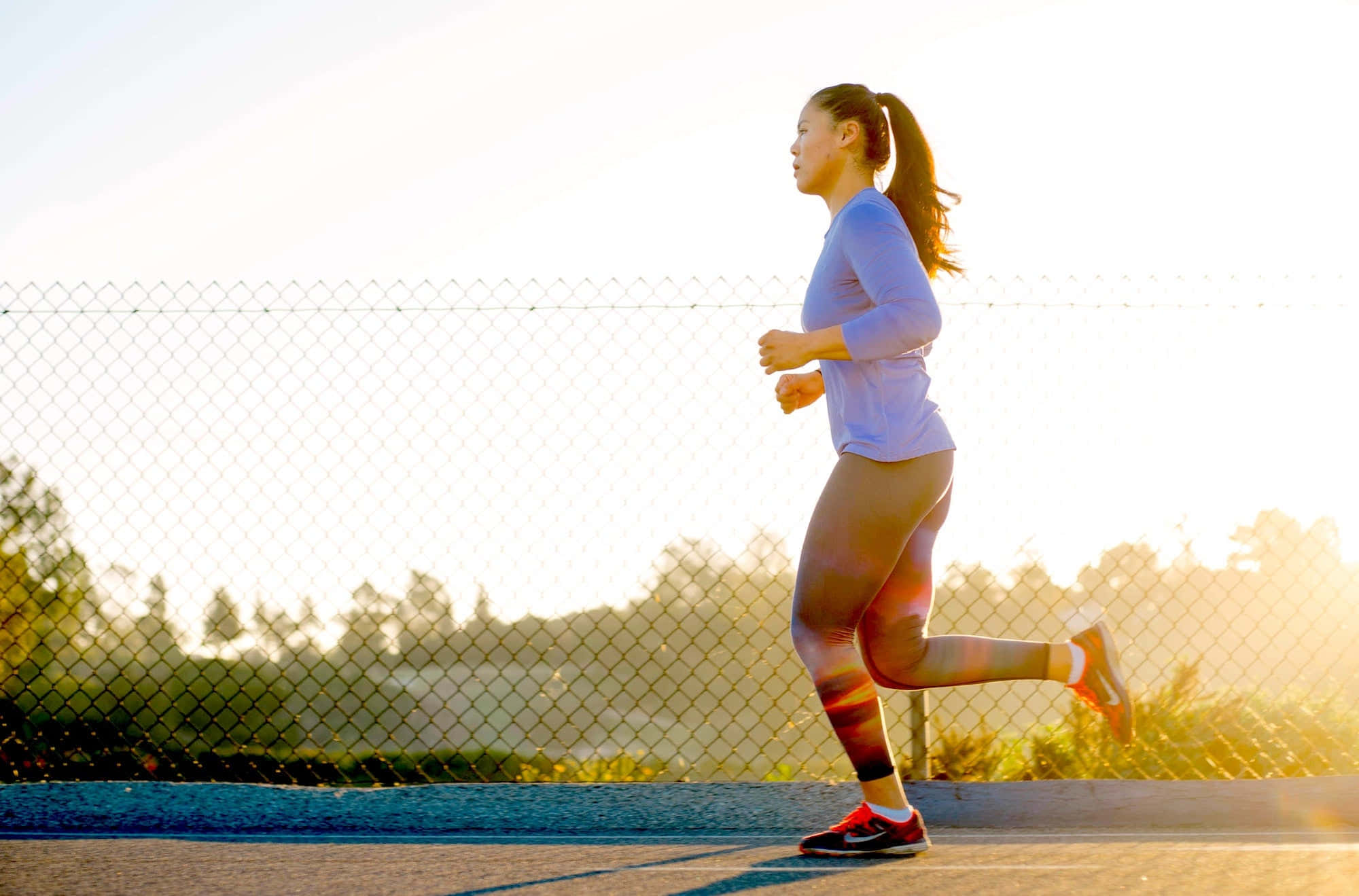 Running 桌布 壁紙 — 一位女士在日出時於戶外跑步，金色陽光在鏈條圍欄旁的鋪砌小徑上投下長長的影子。
