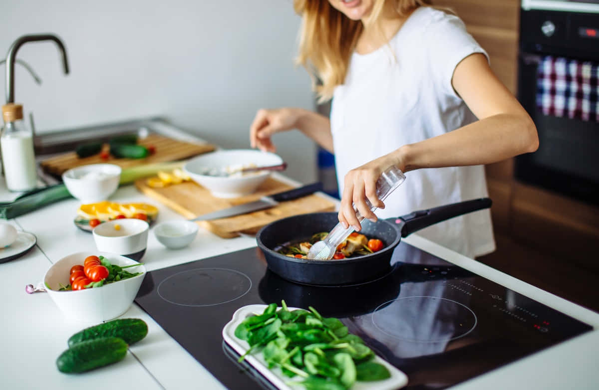 烹飪 桌布 壁紙 — 一位女士在明亮的廚房裡，在現代電磁爐上的煎鍋中為色彩繽紛的蔬菜調味。