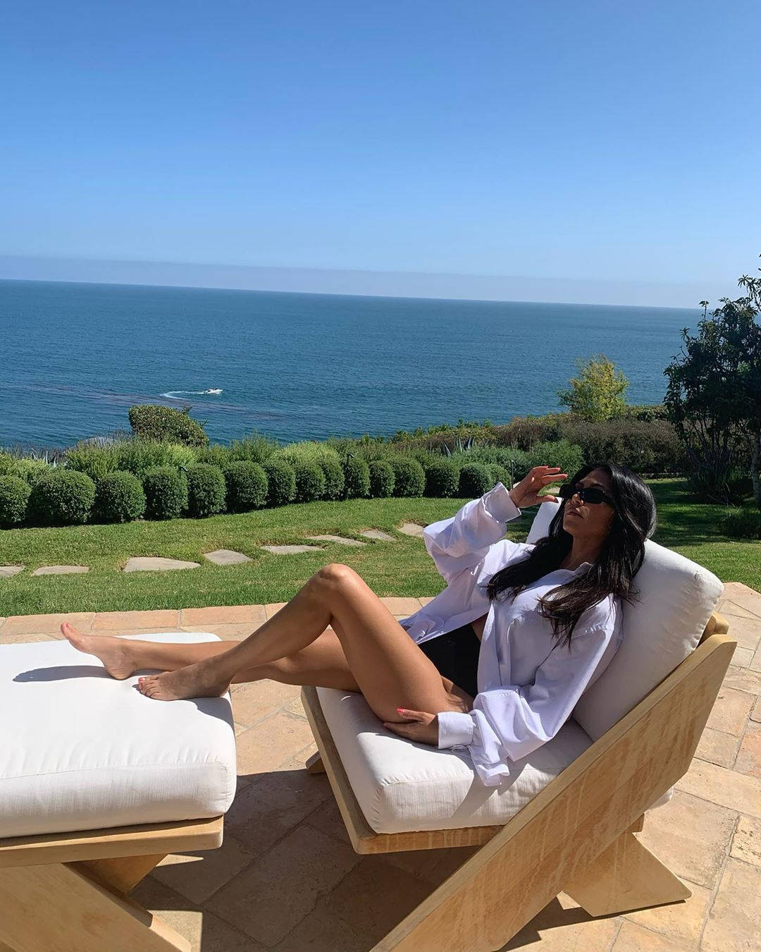 Kourtney 桌布 壁紙 — 一位女士在木椅上休息，身處陽光普照的露台，俯瞰廣闊的藍色海洋和下方修剪整齊的綠植。