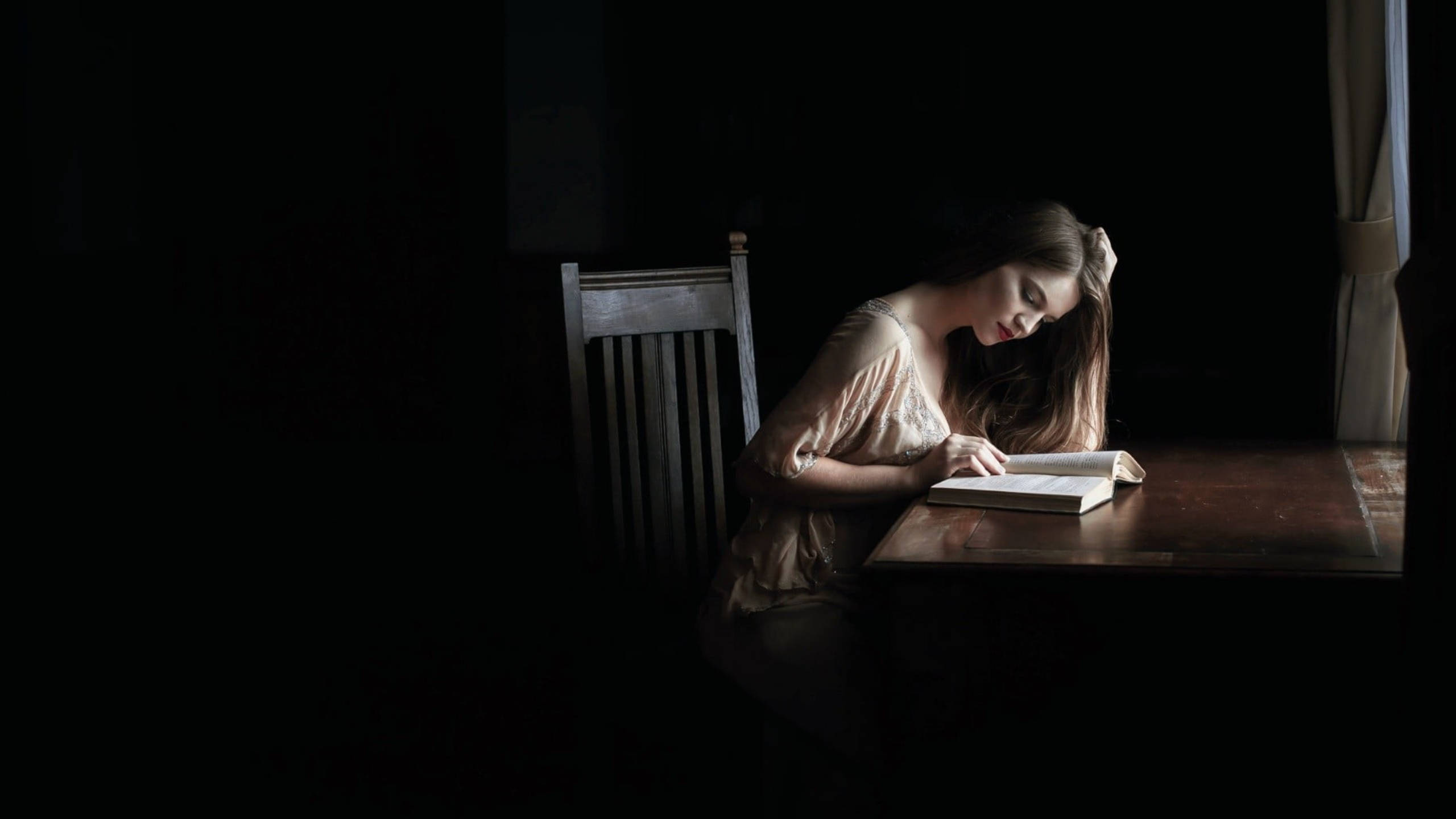 Reading 桌布 壁紙 — 一位女士在柔和光線下，於一個黑暗房間的木桌上閱讀一本書，房間只有一個窗簾。