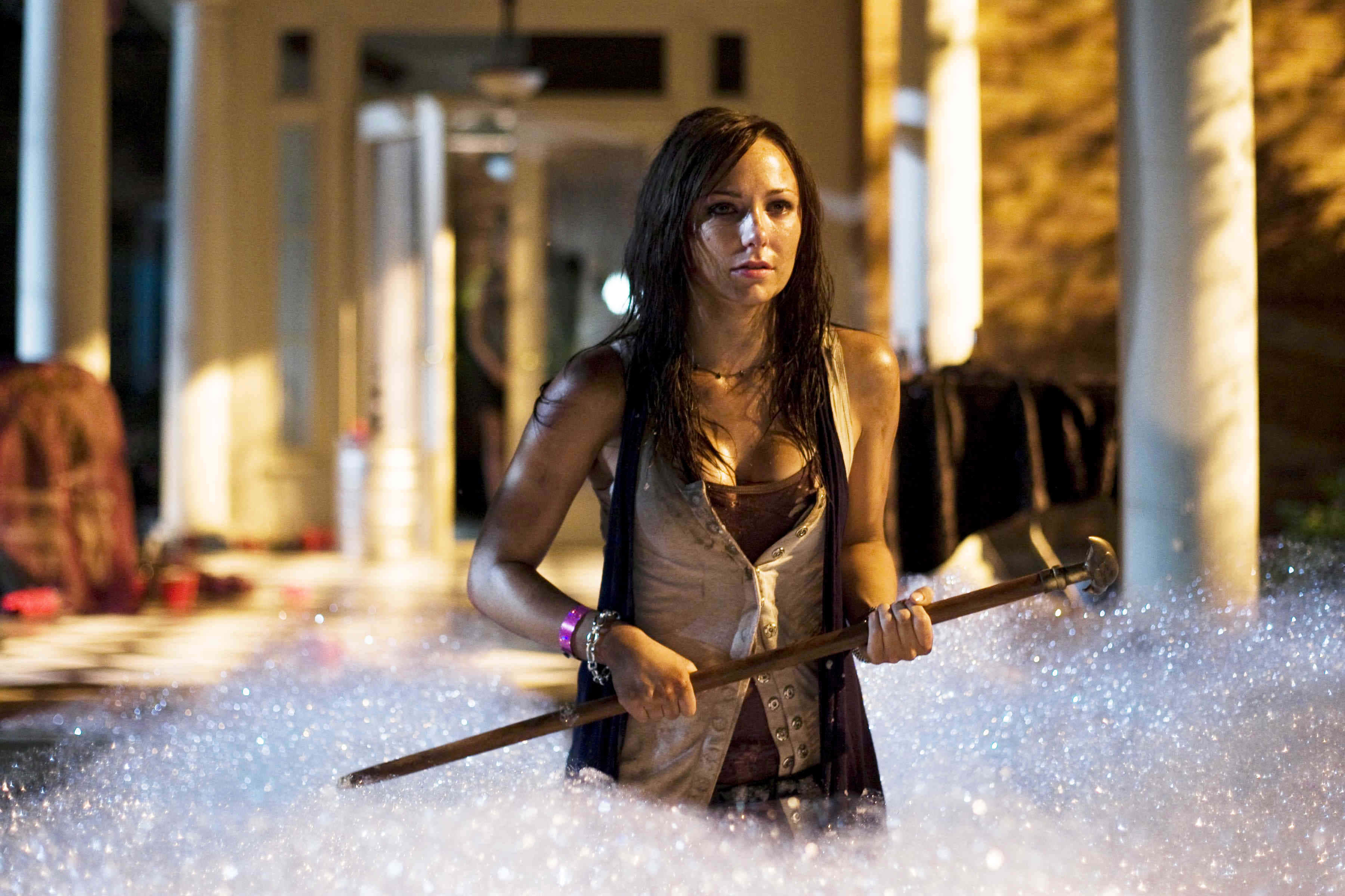 Briana Evigan 桌布 壁紙 — 一位女士在柱廊庭院中，在溫暖的夜燈下，手持杖桿涉水穿過泡沫水。