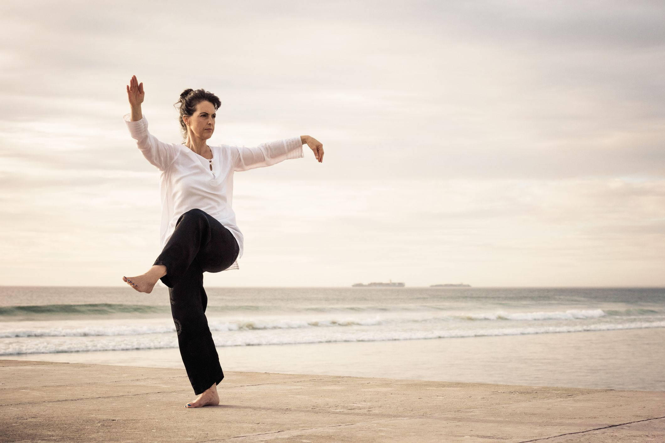 martial arts 桌布 壁紙 — 一位女士在海濱長廊上優雅地平衡著瑜伽姿勢，正值黃金時刻，背景有海浪和遠處的船隻。