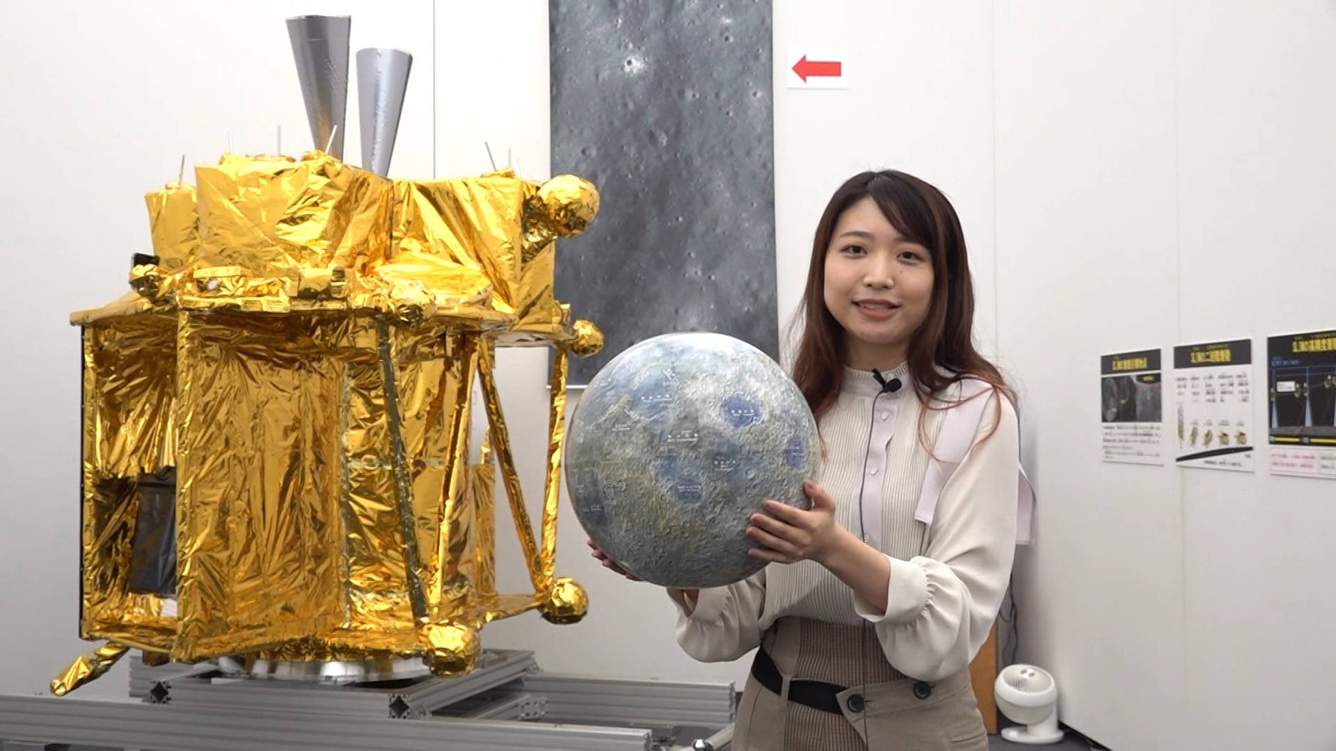 Foil 桌布 壁紙 — 一位女士在潔白的展覽空間中微笑，手持月球模型，旁邊是鍍金箔的太空船複製品。