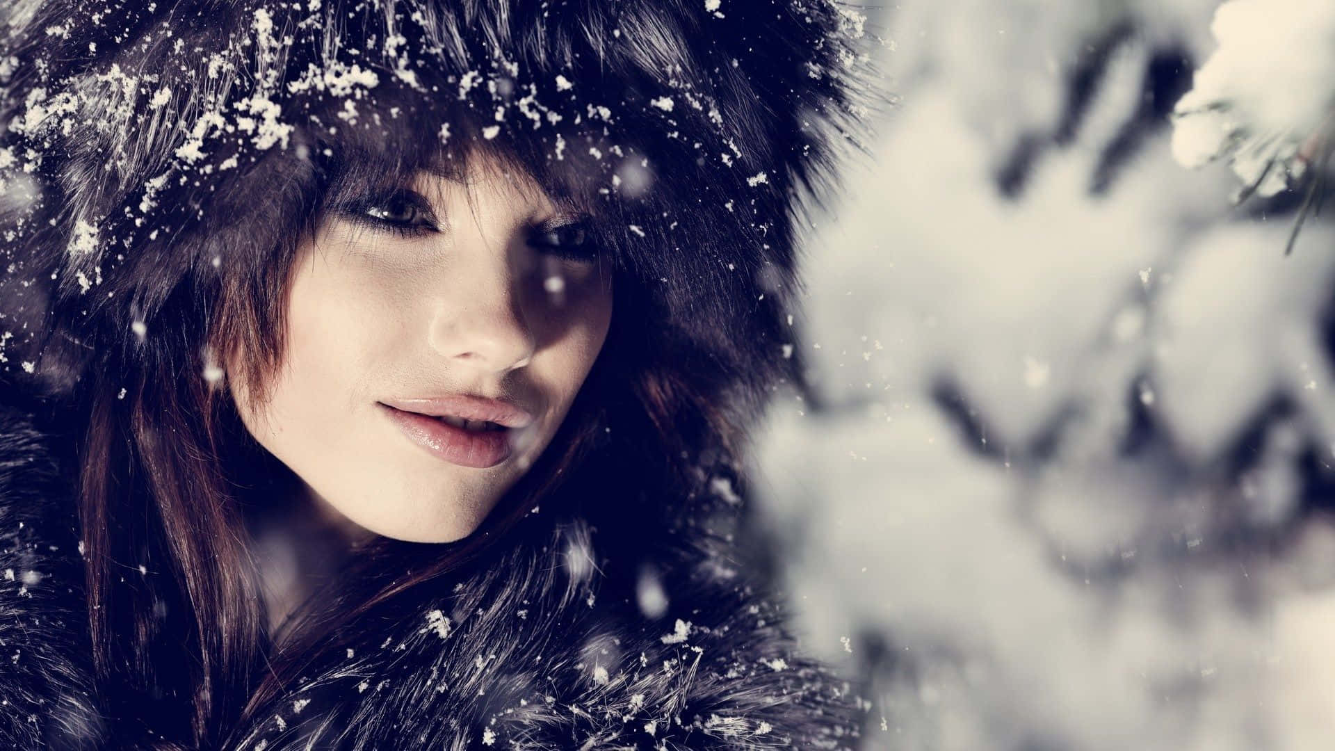 Winter Fashion 桌布 壁紙 — 一位女士在白雪皚皚的冬季森林中，戴著毛皮襯裡的兜帽，雪花輕輕飄落在她的臉龐周圍。