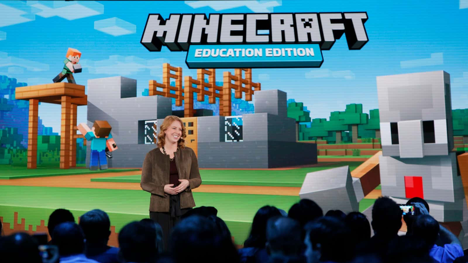 Minecraft Education Edition 桌布 壁紙 — 一位女士在舞台上微笑，背景是Minecraft Education Edition的場景，其中有方塊狀的角色和結構，上方是明亮的藍天。