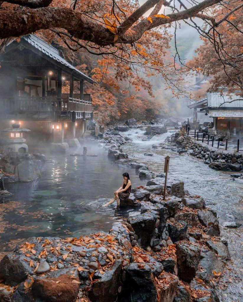 Beautiful Autumn 桌布 壁紙 — 一位女士在蒸氣騰騰的溫泉中放鬆，周圍是秋葉和傳統木建築。