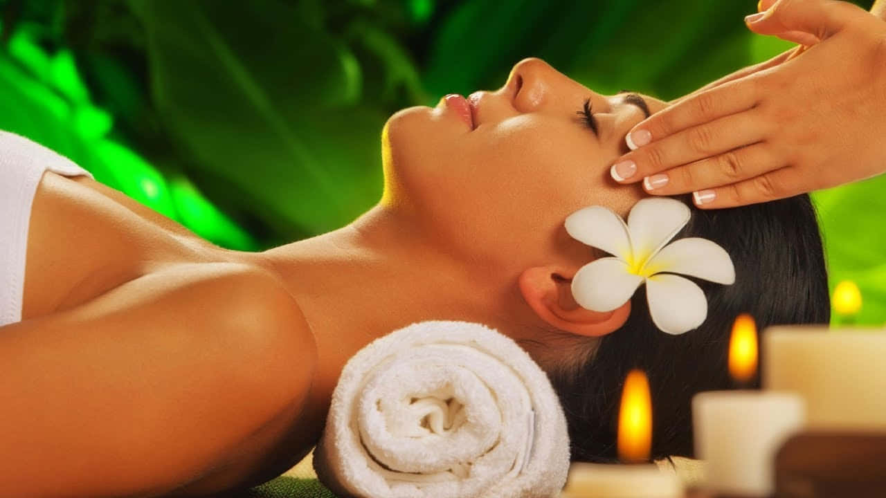 Massage 桌布 壁紙 — 一位女士在閃爍的蠟燭和茂密的綠植中，頭髮上插著雞蛋花，享受著寧靜的臉部按摩。