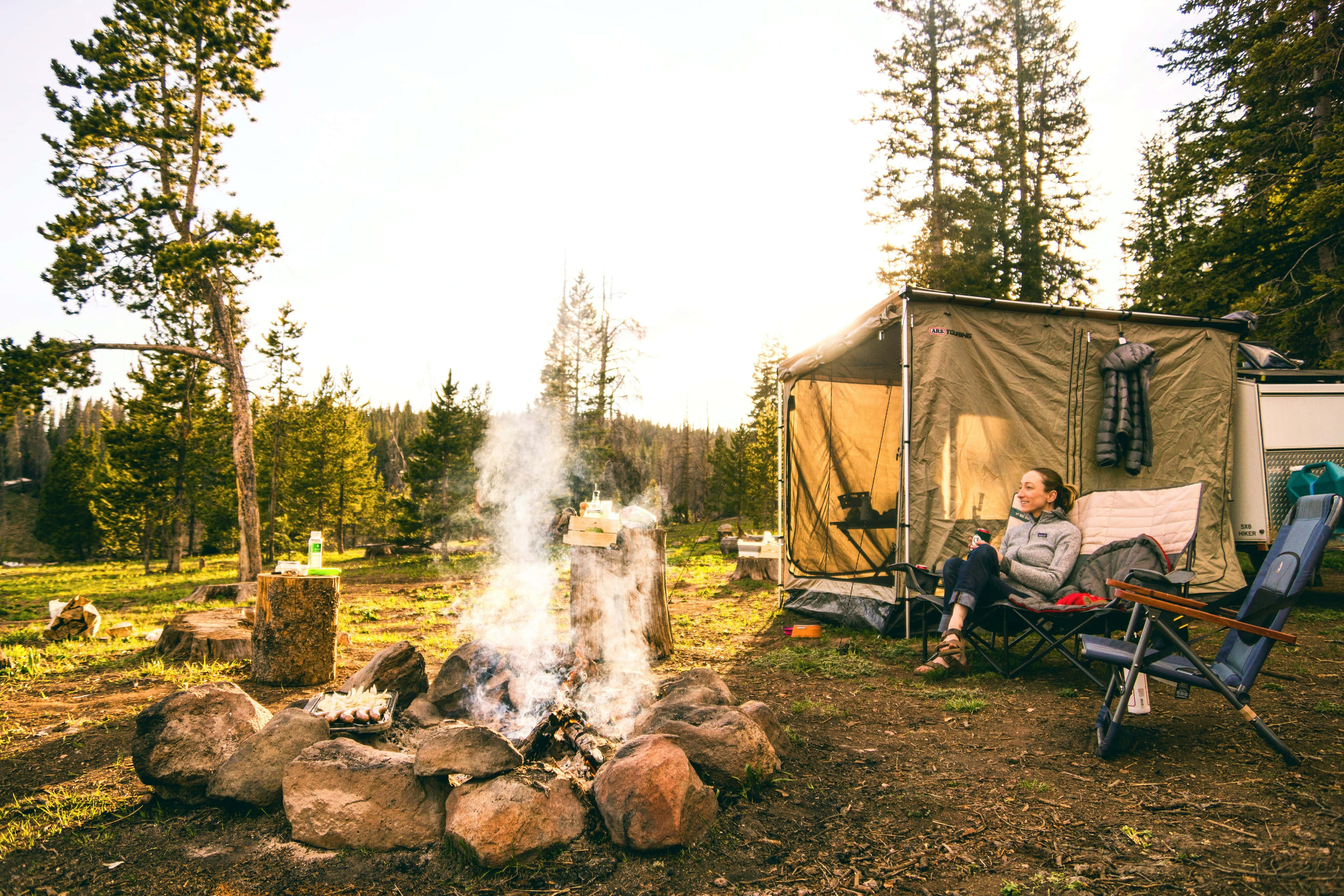 Camping 桌布 壁紙 — 一位女士在陽光普照的松樹林中，在帳篷附近冒煙的營火旁邊的摺疊椅上放鬆。