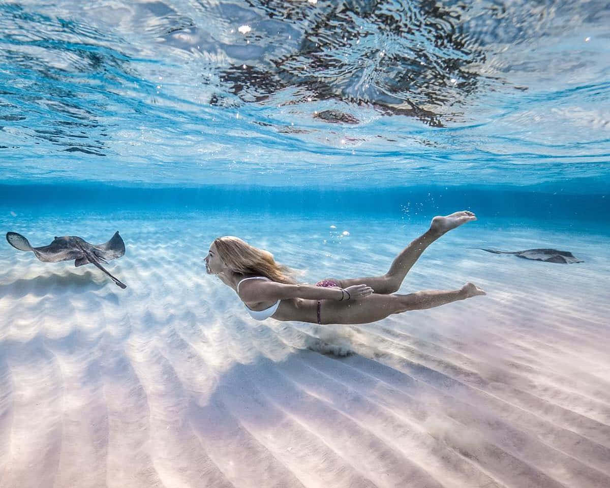 Underwater Ocean 桌布 壁紙 — 一位女士在陽光普照的沙質海底上，優雅地在魟魚旁邊滑行於水中。