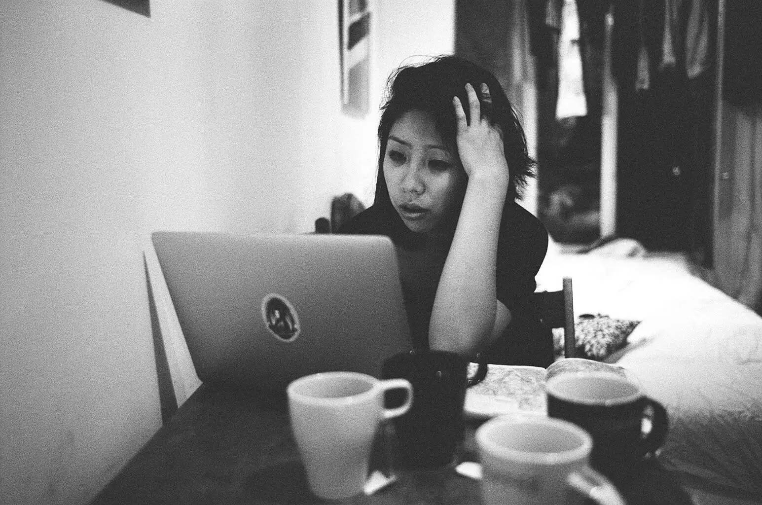 Working 桌布 壁紙 — 一位女士坐在一張雜亂的桌子旁，桌上放著多個杯子，在光線昏暗的房間裡專注地盯著她的筆記型電腦。