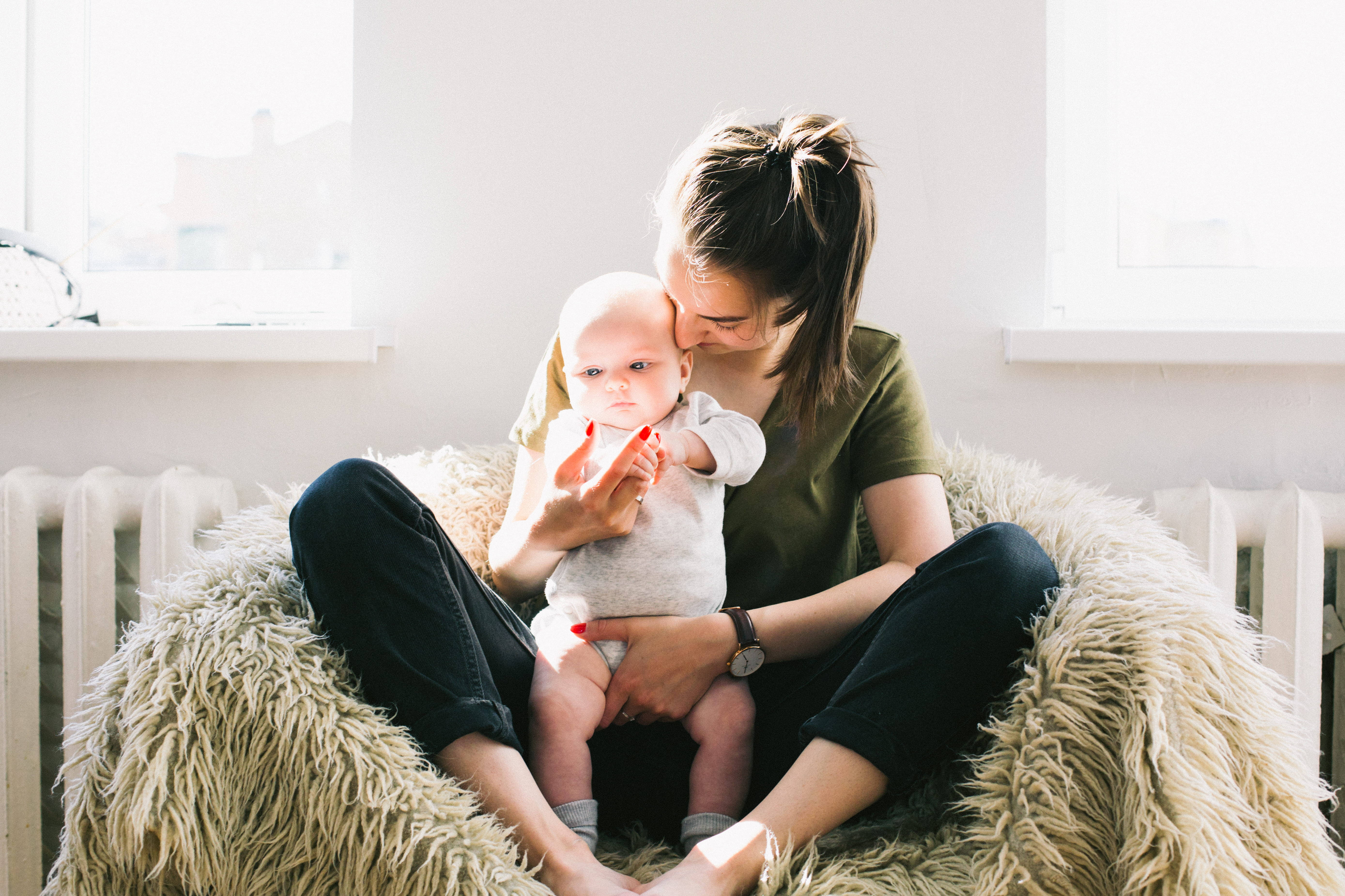Mom 桌布 壁紙 — 一位女士坐在柔軟的米色豆袋椅上，在明亮的陽光房間裡輕輕親吻嬰兒的額頭。