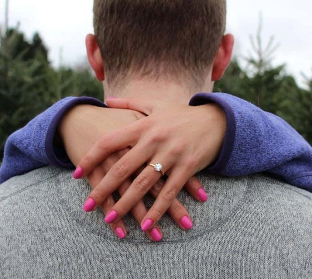 engagement 桌布 壁紙 — 一位女士塗有粉紅色指甲的雙手放在男士的肩膀上，在常綠樹木的背景下展示訂婚戒指。