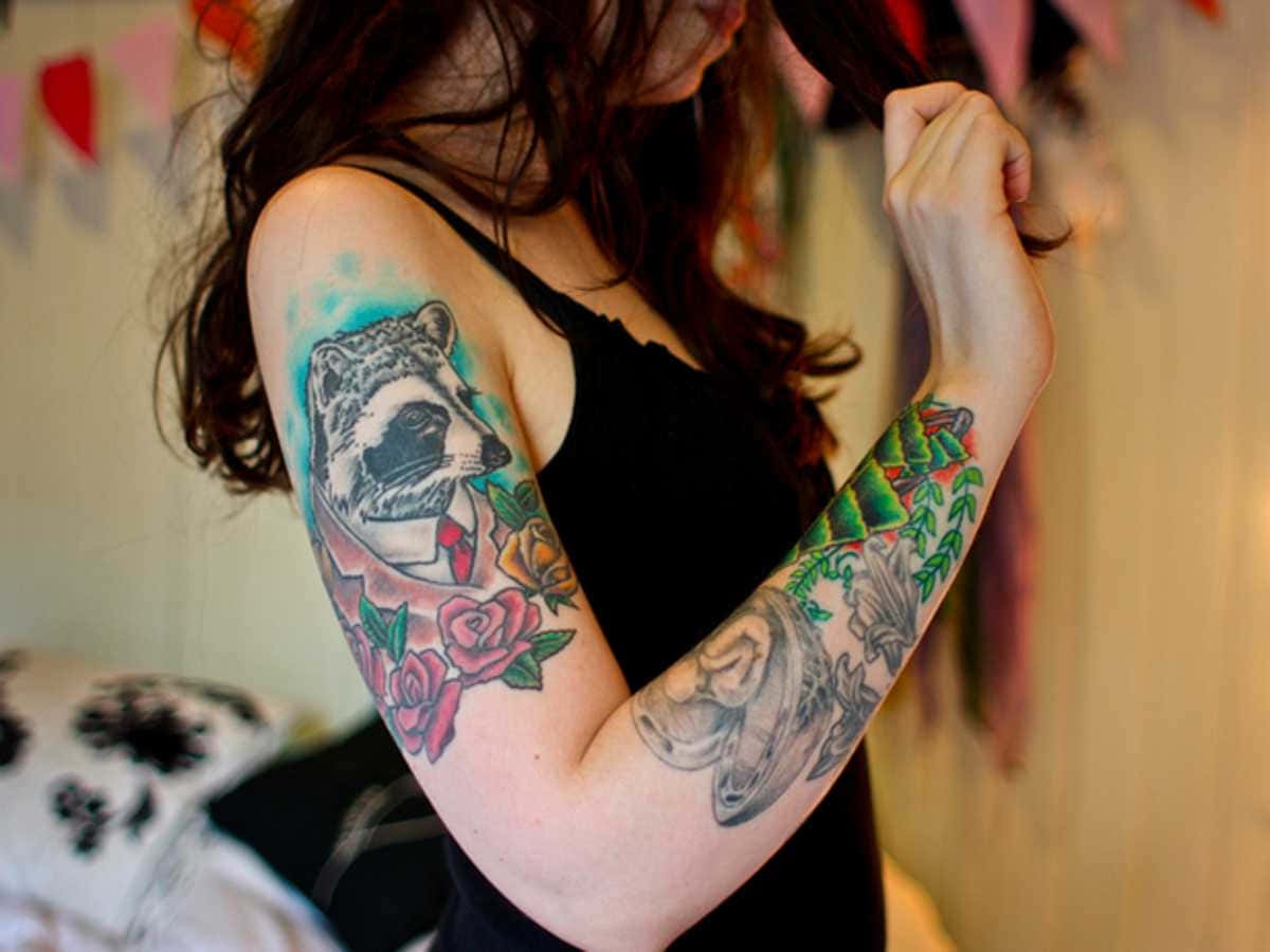 Tattoo Girl 桌布 壁紙 — 一位女士展示手臂上充滿活力的刺青，包括穿著西裝的浣熊、玫瑰和帶有葉子的骷髏頭。